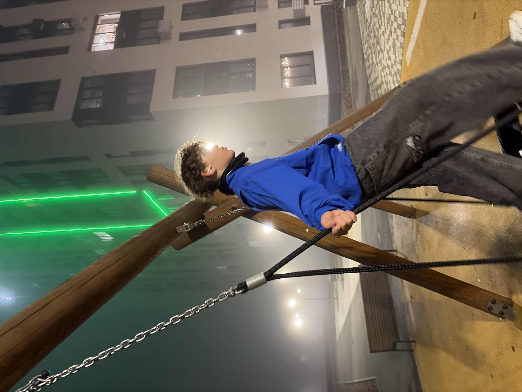 A rotated, low-angle shot depicts a person on a wooden and rope play structure in what appears to be a foggy urban environment. The individual, a young person with short, light hair and a wireless earbud, is wearing a bright blue hooded jacket and dark jeans. They are gripping a black rope element of the structure.
The structure is made of thick wooden beams connected with chains and rope. The background shows a modern apartment building with illuminated windows. There is green light in the environment, potentially from lighting fixtures. The ground surface appears to be paved, with a tiled pattern visible. A bench is also partially visible on the ground.
Based on the available information, it is not possible to definitively confirm the location as Izhevsk, Russia.