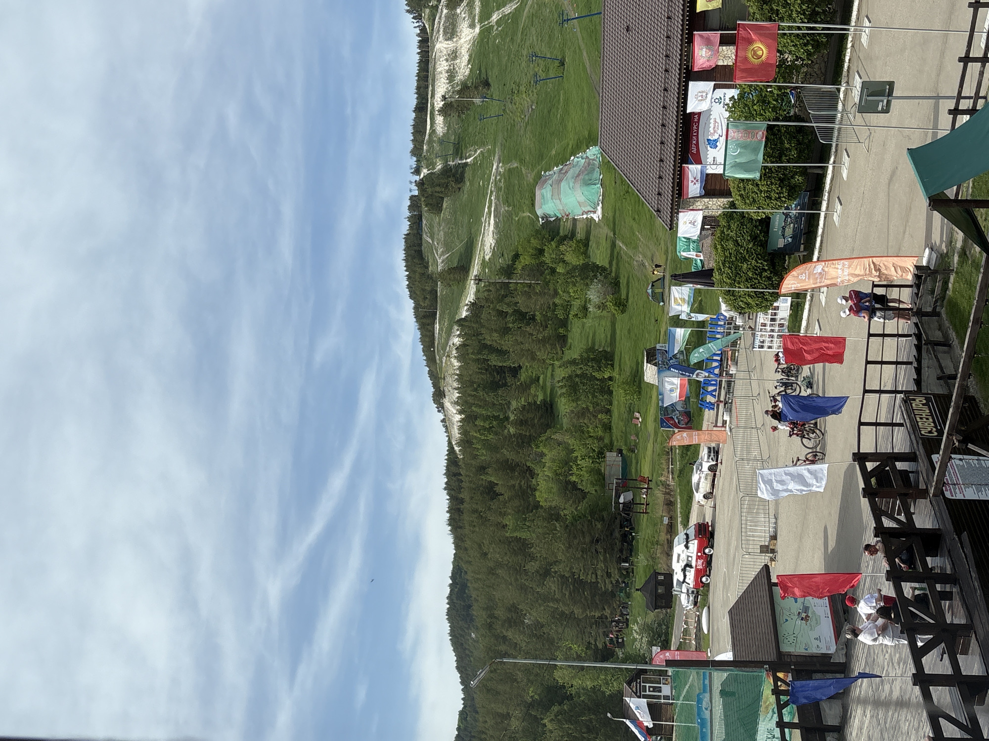 The image depicts a scene at the Khvalynsk Ski Resort in Khvalynsk, Russia. It appears to be a sunny day with a blue sky with wispy clouds. A grassy hill is visible on the left side of the frame with ski lifts and trails. The right side features a building with a brown roof, adorned with various flags representing different nations or organizations. A paved area in front of the building shows parked cars, cyclists, and pedestrians. A wooden structure, possibly a deck or platform, is visible in the foreground with people and objects on it.