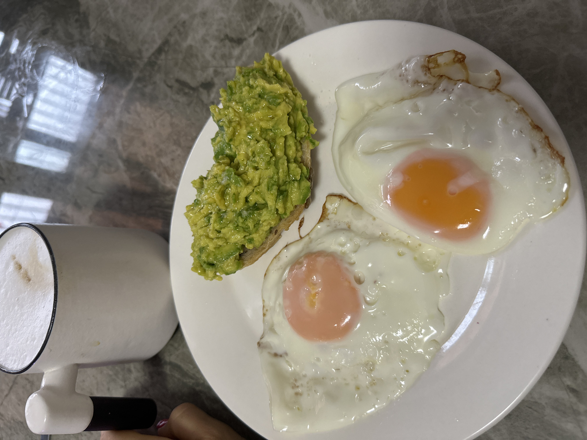 The image shows a plate of food on a marble-patterned surface. The plate contains two fried eggs and a piece of avocado toast. A white mug with a black handle sits to the left of the plate. A person's hand is partially visible in the lower left corner of the image.
There is no location context within the image, therefore the location "Bac Nha Trang Ward, Vietnam" cannot be confirmed.