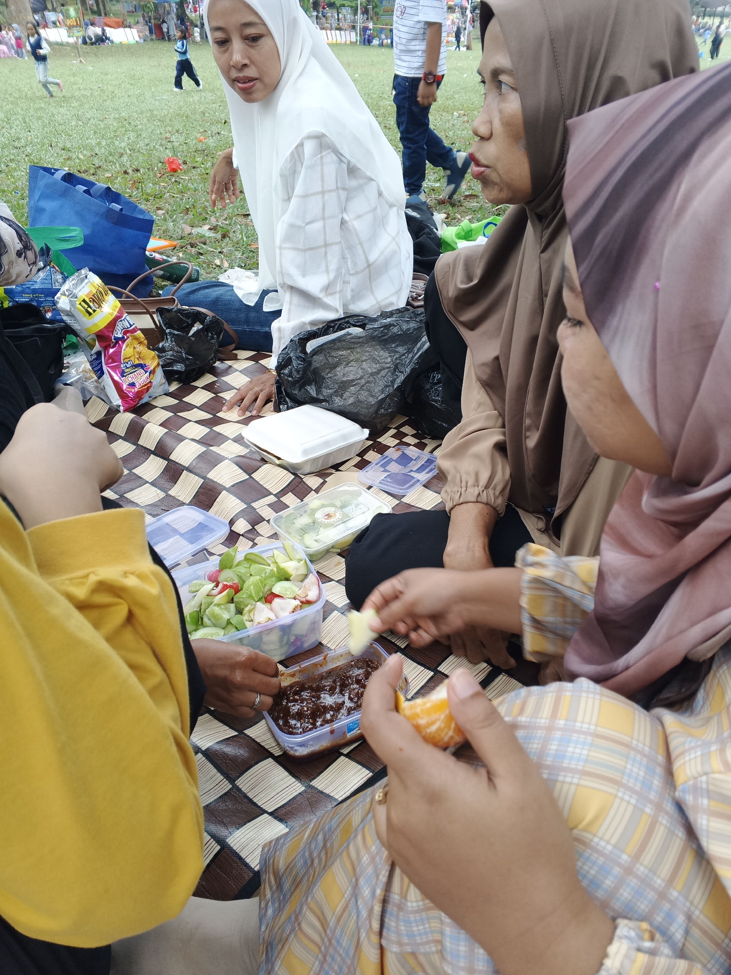 A group of women and children are picnicking on a checkered mat outdoors. The women are wearing hijabs, one in white, one in brown, and another in a pale pink. They are seated on the mat, with various food containers and snacks spread out. One woman in a yellow jacket is holding a piece of orange. In the background, people are visible on a grassy field, and there are tents and structures that suggest a park or event location, possibly in the city of Perbawati, Indonesia.