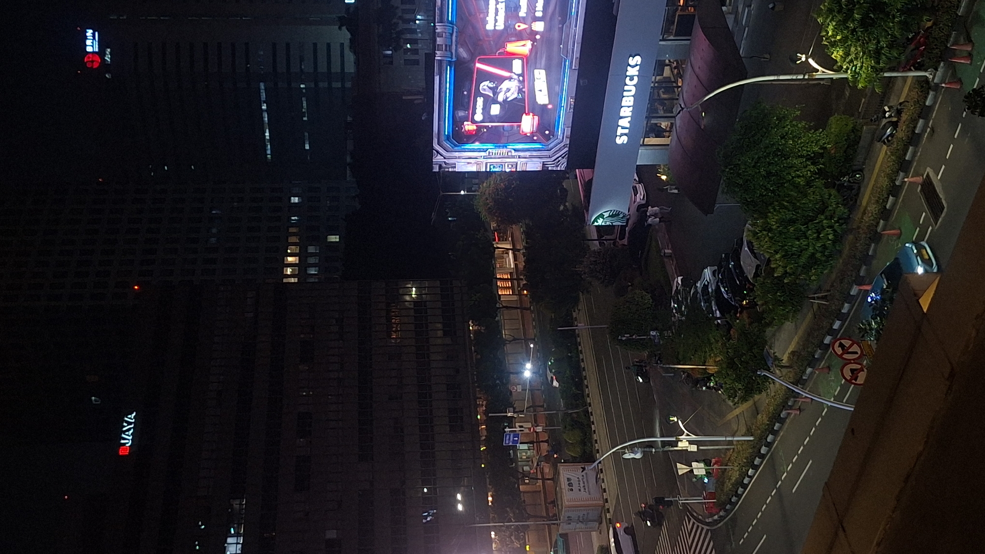 The image shows a nighttime view of Central Jakarta, Indonesia. Prominently visible is a Starbucks store adjacent to a large digital display showing an advertisement for a mobile game. A multilane road with traffic lights is visible, with cars and motorcycles on the road. Tall buildings are visible, including one with the word "JAYA" illuminated vertically in red. The scene is lit by streetlights, building lights, and the bright digital display.