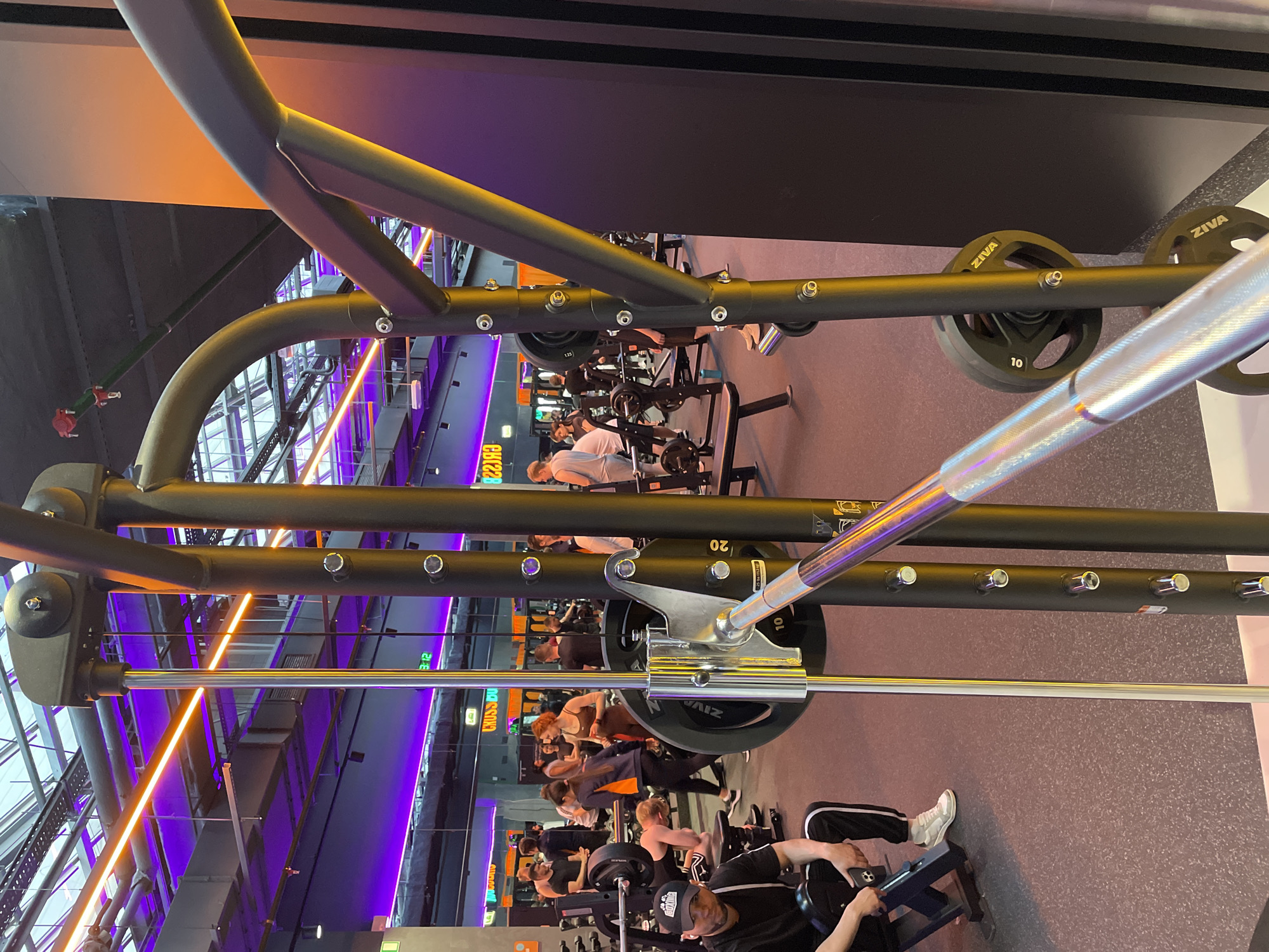 The image depicts a gym interior. In the foreground, a Smith machine with weights is visible. People are actively engaged in exercising on various machines in the background. The gym is brightly lit with a combination of orange and purple linear lighting. Kazan, Russia, is assumed to be the location based on the description.