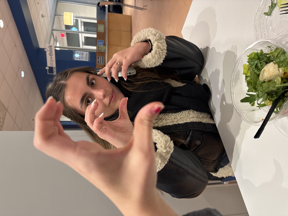 A woman wearing a black jacket with a fur collar is talking on a cell phone. Her left hand is raised towards the viewer. On a white table next to her is a plastic container with a salad. A fork is visible in a second, similar container. The image is rotated counter-clockwise. The background shows what appears to be a cafeteria or seating area.