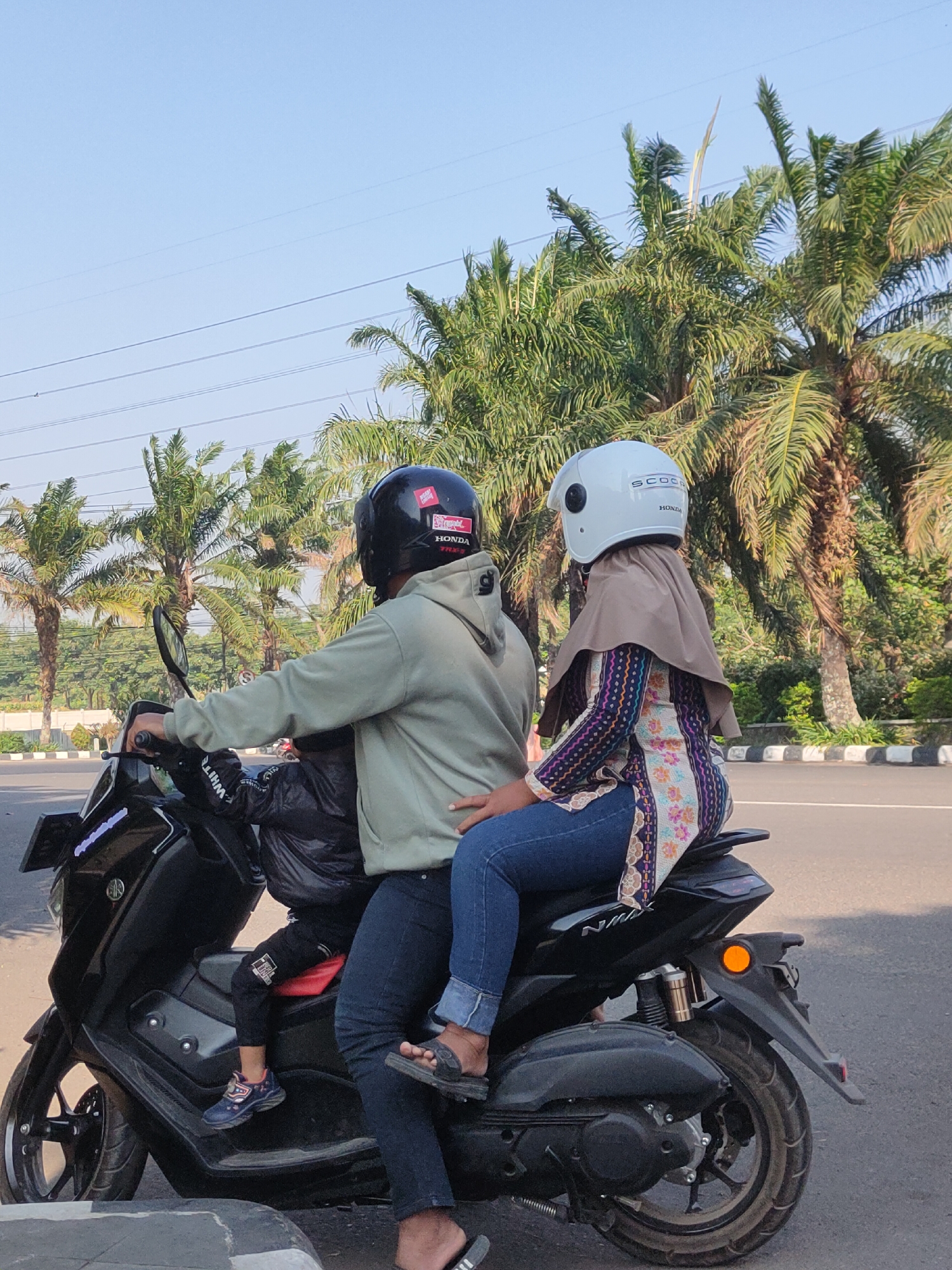 The image depicts three individuals riding a black scooter on a paved road, likely in Ngasem, Indonesia, given the context. The scooter is moving forward, and the individuals are facing the same direction.
In the front is a person wearing a black helmet with a red sticker on it, a light green hooded sweatshirt, and dark jeans. They are operating the scooter. Behind them, a younger person is seated, wearing a black jacket and dark pants with a red cushion visible. Seated behind the driver is another individual wearing a white helmet, a tan hijab, and a long-sleeved patterned shirt with blue jeans. Their left arm is around the driver.
The environment appears to be a suburban or roadside area. Tall palm trees line the right side of the road, with dense green foliage behind them. Overhead, power lines stretch across a clear blue sky. The road has a striped white and black border, suggesting it's a designated lane or shoulder. The lighting indicates it is daytime, likely late morning or early afternoon, with clear visibility.
