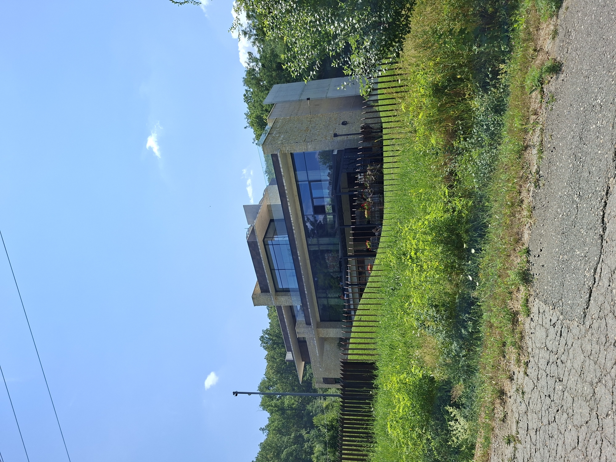 In the city of Condrița, Moldova, a modern, multi-story house is viewed from a slightly elevated position across a cracked asphalt road and a metal fence. The house, with its angular design, features stone and concrete facades, large glass windows, and cantilevered sections. A balcony with glass railings is visible on the upper level. The surrounding environment includes lush green grass and dense trees on a hillside behind the property. The sky is clear blue with a few wispy clouds. A tall pole with a security camera is positioned to the left of the house. The scene appears to be captured during daylight.