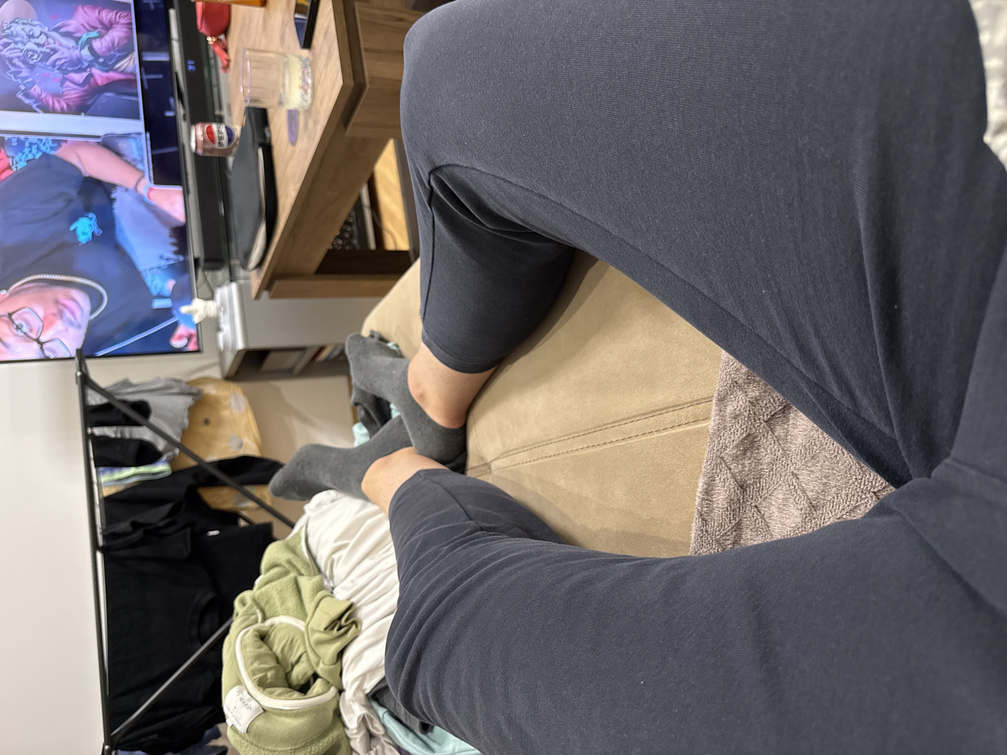 A high-angle, indoor shot depicts a person wearing dark blue sweatpants and gray socks, sitting on a tan couch. To the left, a television displays a video of people. A wooden table is partially visible above the person's leg, holding items including a can and a glass. A black metal clothing rack filled with clothes is situated to the left and below the person's feet. The location and city of Lovech, Bulgaria are not discernible from the visual information in the image.