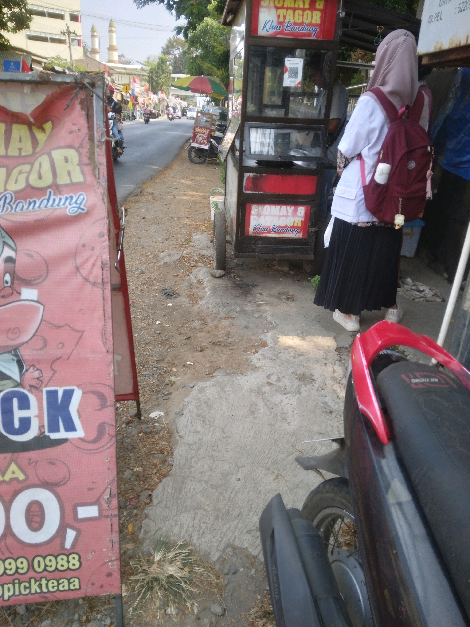 The image depicts a street scene in Desa Pamijen, Kecamatan Sokaraja, Indonesia. On the left, a large red sign with cartoon characters and text advertising "AY GOR Bandung" is visible. To the right of the sign is a street with several motorcycles and a food cart. The food cart has red trim and signs indicating "SIOMAY & BATAGOR Khas Bandung." A young woman wearing a hijab, white shirt, and black pleated skirt is standing in front of the food cart, carrying a maroon backpack. She is wearing white sneakers. A partially visible motorcycle is in the foreground on the right. The environment appears to be a typical street with a paved surface and dirt/gravel areas. In the background, there are buildings, trees, and a mosque with two minarets. The sky is overcast.