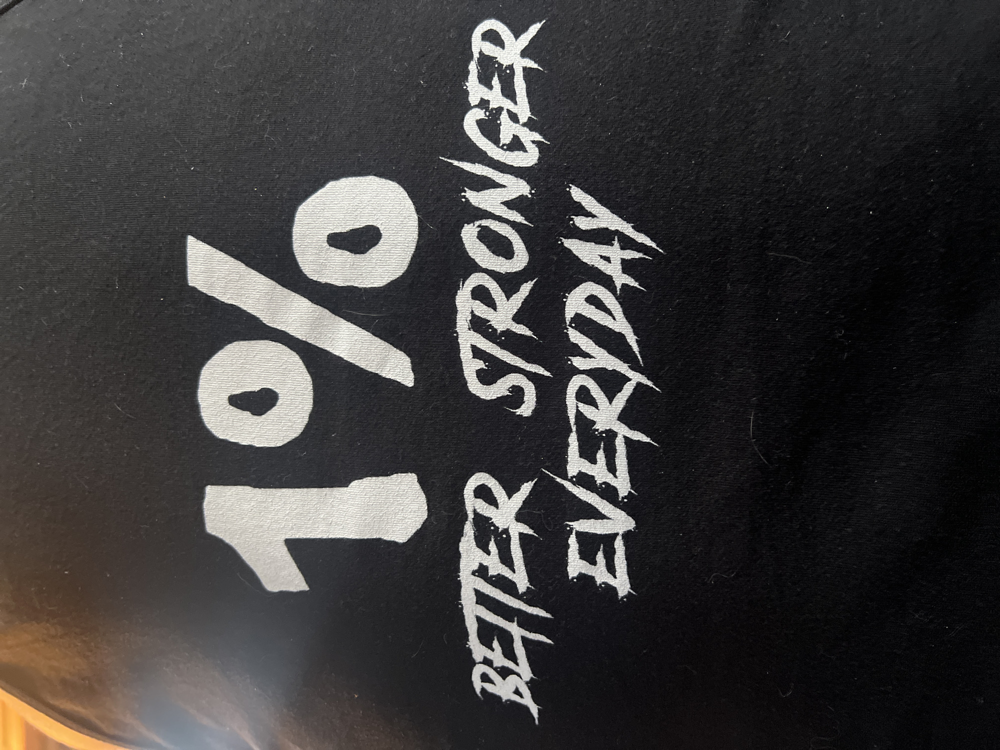 The image shows a close-up of a black fabric, likely a shirt, with white text printed on it. The text reads "1% BETTER STRONGER EVERYDAY," stacked vertically. The words "BETTER STRONGER EVERYDAY" are in a jagged, almost aggressive font. There is no indication of the image being in City of Newburgh, United States of America.