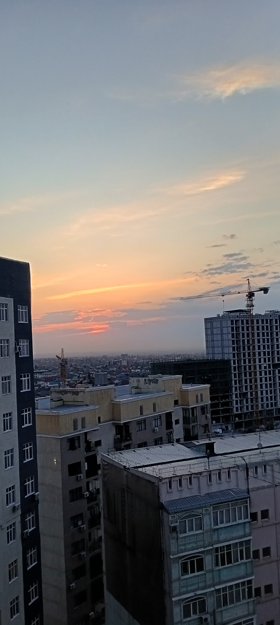 The image depicts a cityscape at sunset, likely in Bishkek City, Kyrgyzstan, given the architectural style and distant view of buildings and low-rise structures. The sky is a gradient of pale blue at the top, transitioning to soft orange and pink hues near the horizon, indicating the setting sun. Wispy clouds are scattered across the sky.
Dominating the foreground and midground are several multi-story buildings, indicative of an urban environment. The building on the far left is tall and narrow, with a dark grey facade and numerous windows arranged vertically. Adjacent to it are other apartment buildings, varying in height and architectural style, some with lighter colored facades and balconies. One building appears to be under construction, with a concrete frame and scaffolding visible.
In the midground, a construction crane is prominently positioned, its boom extended horizontally across the frame. Further in the distance, a dense collection of lower-rise buildings, characteristic of residential areas, stretches towards the horizon. The overall impression is of a developed urban area with ongoing construction. No people are visible in the frame. The lighting suggests it is either early morning or late evening.