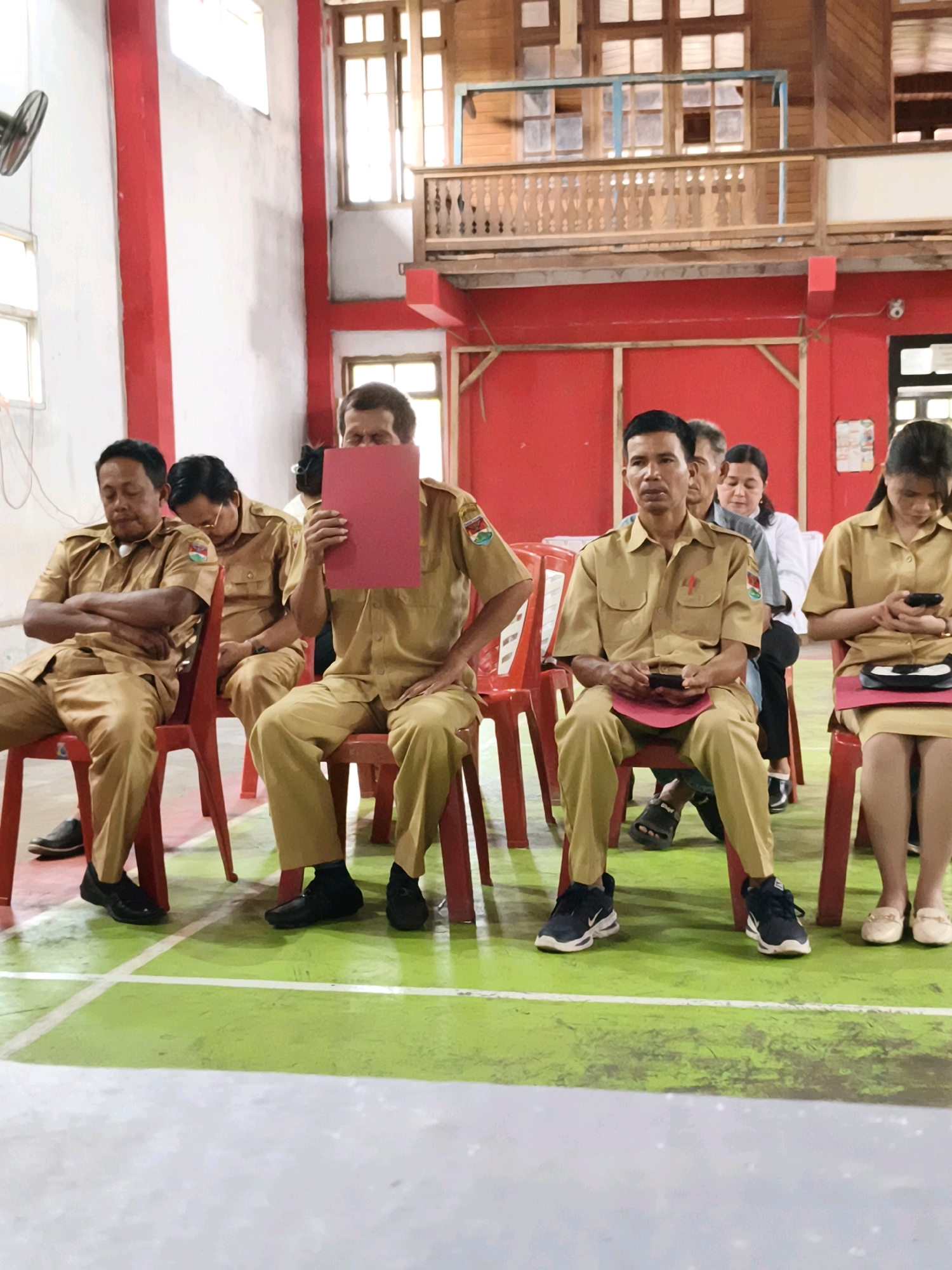 In Tombatu Satu, Indonesia, an indoor scene captures a group of people, likely civil servants, seated on red plastic chairs in what appears to be a community hall or school gymnasium. The flooring is a light green with white court markings. The walls are painted white and bright red, with exposed wooden beams and window frames visible in the background, suggesting a rustic architectural style.
In the foreground, four men and two women are seated. The men are all wearing matching tan uniforms with dark epaulets and patches on the sleeves. One man in the center is holding up a red folder, obscuring his face. Another man to his left is holding a dark object, possibly a phone, and a red folder on his lap. The man on the far left is leaning back with his arms crossed, his eyes closed.
To the right of the men, two women are seated. The woman in the foreground is wearing a tan uniform similar to the men's, holding a dark mobile phone in her hands. The woman behind her is wearing a white top and dark pants, with her head slightly tilted down.
The overall impression is one of a formal gathering or meeting, possibly during a break or presentation. The lighting is bright and diffused, indicating an indoor setting with natural light sources. The scene is captured from a low-angle perspective, focusing on the seated individuals.