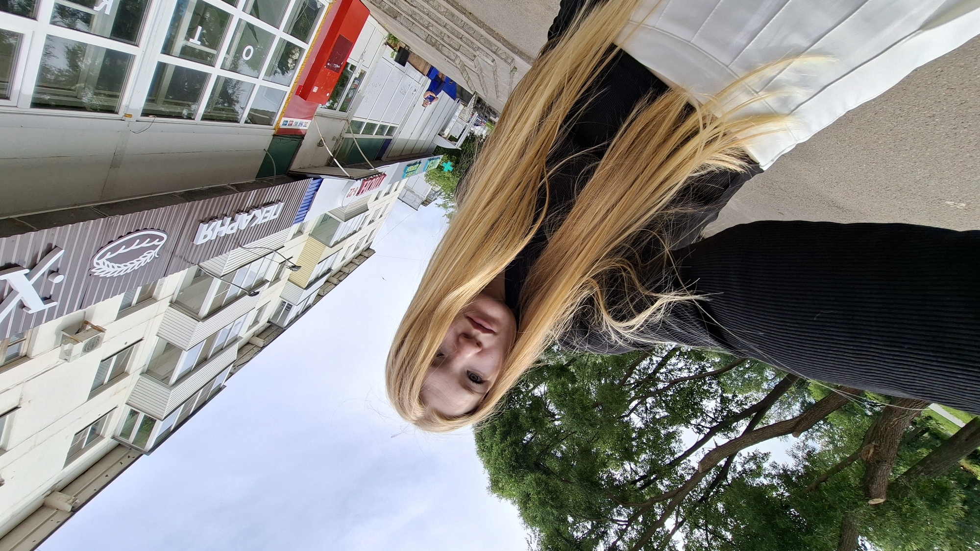 A young woman with long blonde hair stands outside a building in Chaikovsky, Russia. She is wearing a black top and a white skirt. The building behind her has a sign that reads "ПЕКАРНЯ," which translates to "Bakery." There are trees and a cloudy sky in the background. The image is taken from a low angle, looking up at the woman and the building.