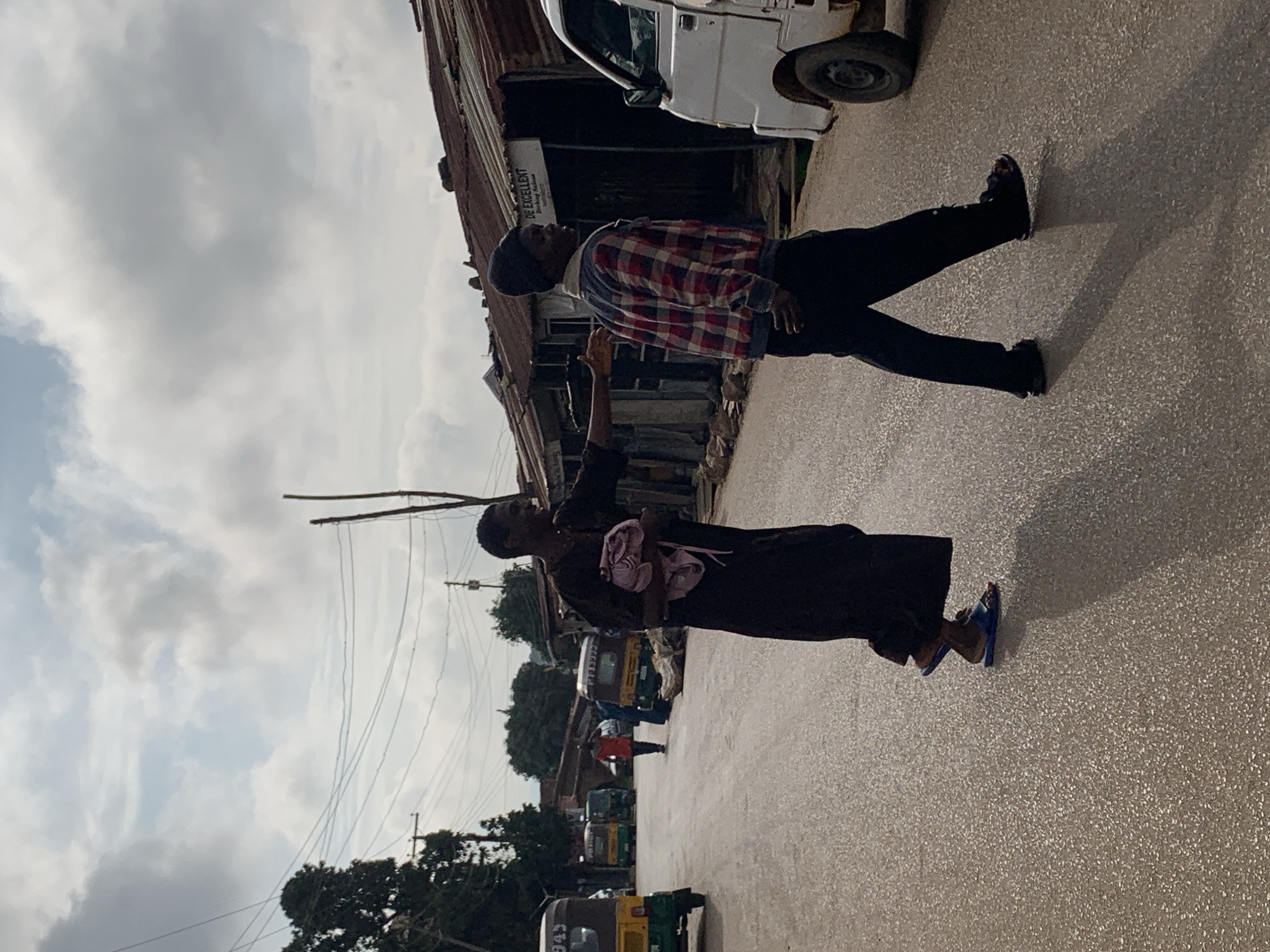 In Jos, Nigeria, a street scene unfolds with two individuals crossing a paved road. The person on the left, wearing a dark, flowing garment and blue flip-flops, carries a bundle of pink fabric. They appear to be reaching out towards the other person. The second individual, walking to the right, is clad in a red and black plaid jacket, a dark beanie, and dark pants, with dark footwear. Behind them, to the right, a white utility vehicle is parked on the side of the road. Further in the background, to the left, several auto-rickshaws are visible, one light yellow and two darker colors. The street is lined with buildings with corrugated metal roofs, one of which has a sign reading "DE EXCELLENT." The sky above is partly cloudy, with a few trees and utility poles also present in the environment.
