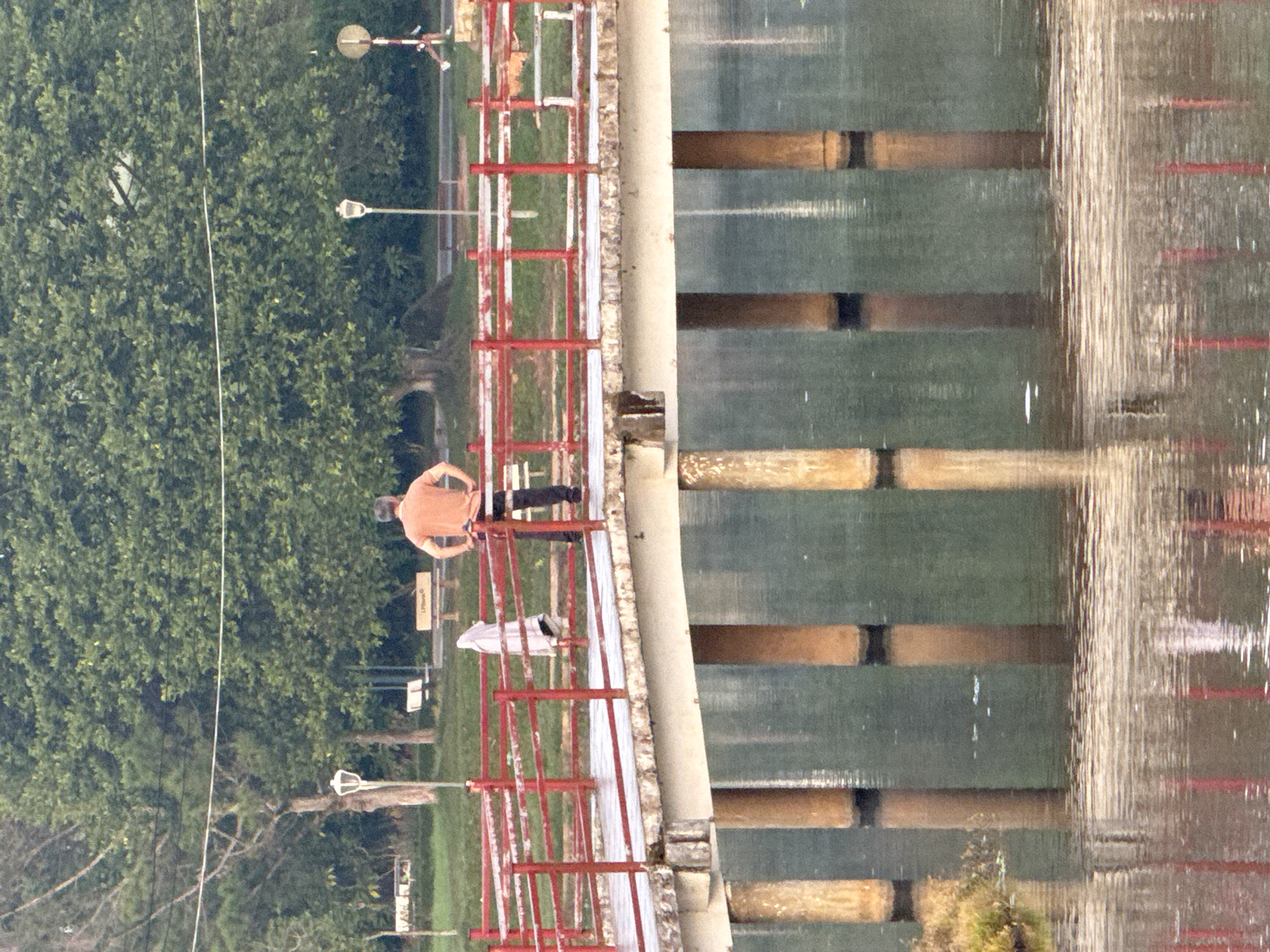 A man stands with his hands on his hips on a bridge with red railings, overlooking a body of water. He is wearing an orange t-shirt and dark pants. A white garment, possibly a towel or shirt, is draped over the railing to his left. The bridge, supported by thick wooden pillars, spans across the water. The surroundings include lush green trees, grass, and benches in the background. The scene appears to be in a park or recreational area in Lâm Viên - Đà Lạt, Vietnam, given the landscape and likely climate. The water's surface reflects the bridge and the trees, indicating a calm day.