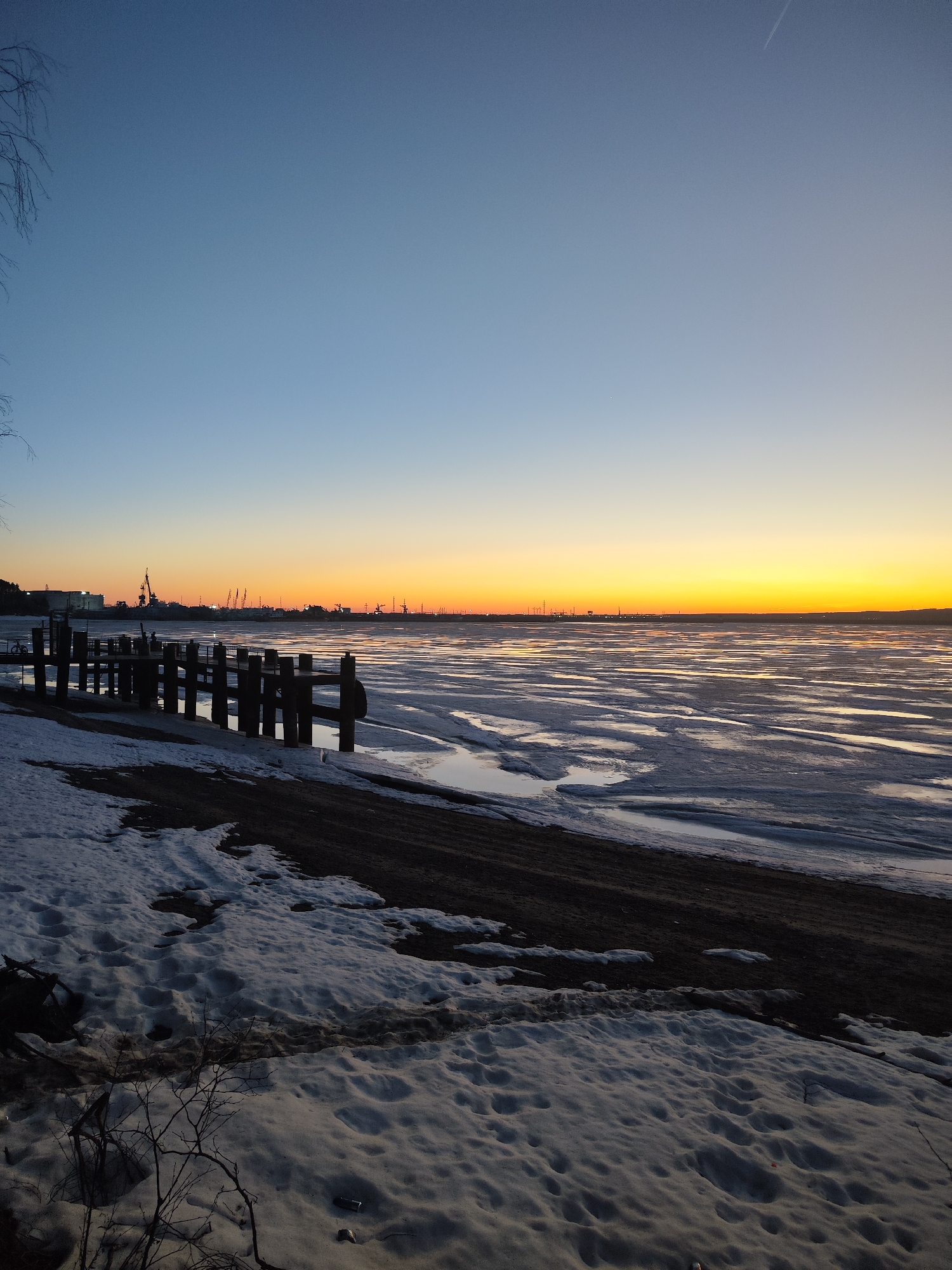 The image shows a frozen body of water, possibly a river or a bay, near a shoreline covered with snow. A wooden pier extends into the water. In the distance, a cityscape silhouette is visible against the sunset sky, which transitions from blue to orange. The scene appears to be in an industrial or urban area.