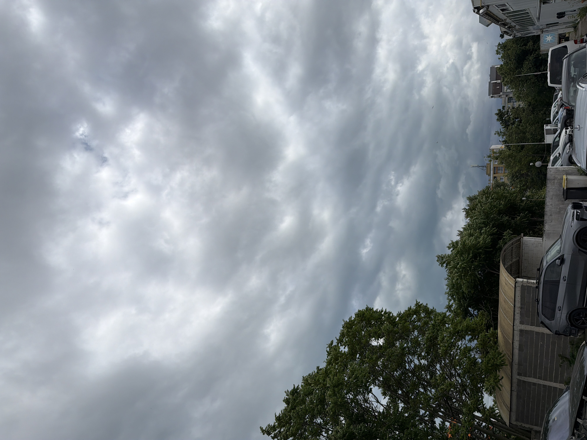 The image shows a cloudy sky over a parking lot in Burgas, Bulgaria. Several cars are parked in the lot, including a grey SUV and a white van. There are also some trees and buildings visible in the background. The sky is filled with heavy, grey clouds, suggesting that it may rain soon.