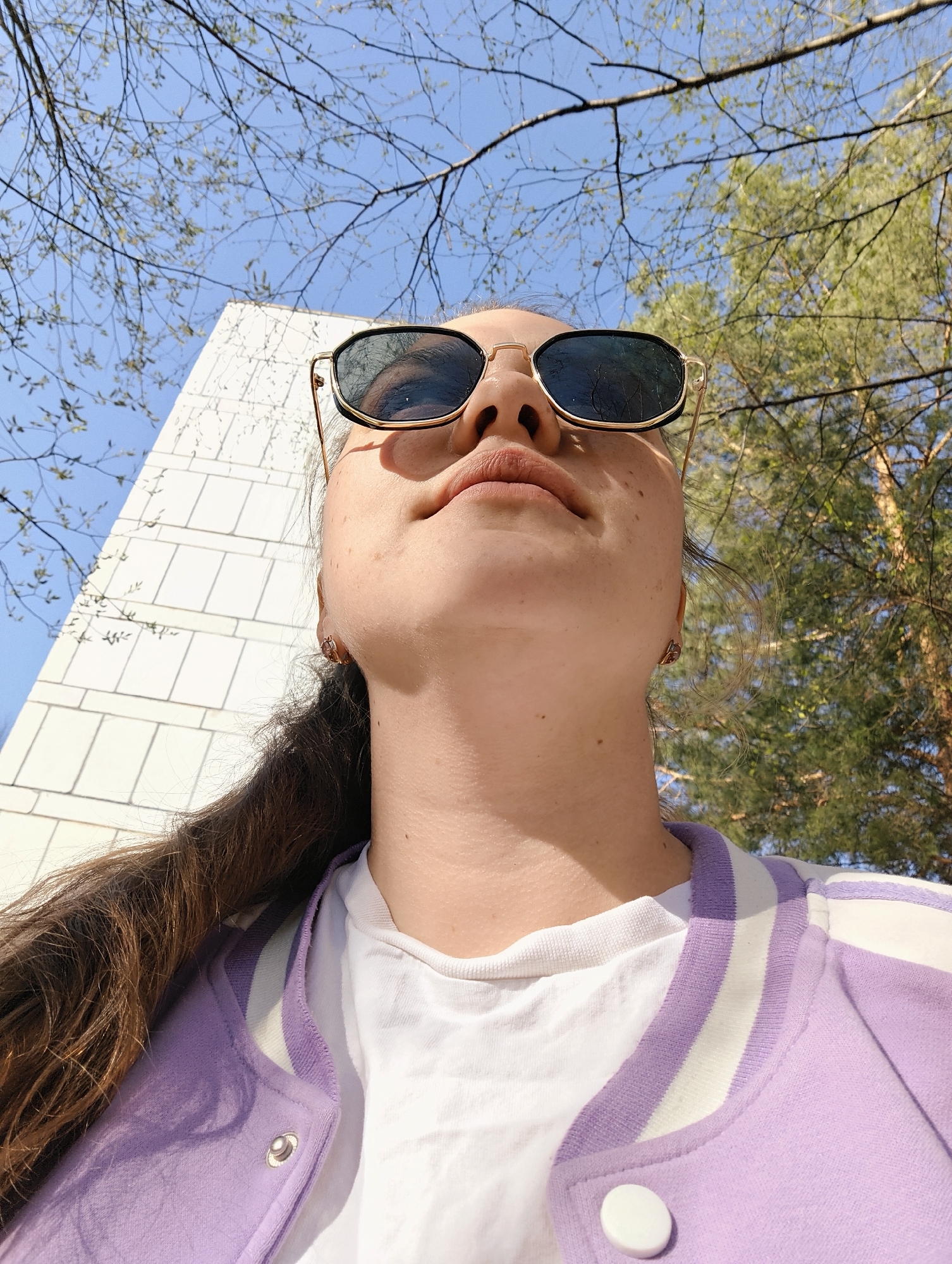 The photograph depicts a person wearing sunglasses, a white t-shirt, and a purple and white jacket. The view is from a low angle, looking upwards towards the person's face. Behind the person, there is a building with white rectangular panels and trees with budding leaves. The sky is visible in the background. Based on the appearance of the building and the vegetation, it is likely that this photograph was taken in the city of Chaikovsky, Russia, but that could not be confirmed.