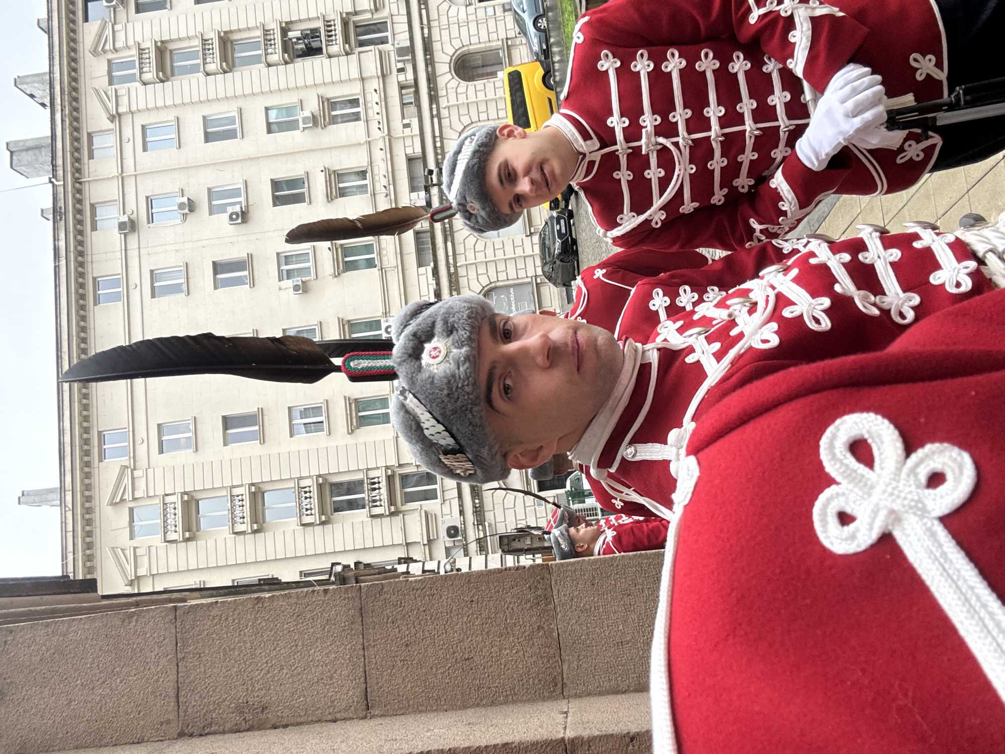 Two uniformed guards stand on a stone step in Sofia, Bulgaria. Both wear gray fur hats with green and red bands, and large black feathers. Their bright red uniforms feature intricate white knotted detailing. Behind them, a large white building with repetitive window patterns is visible. A yellow vehicle is partially visible in the background.