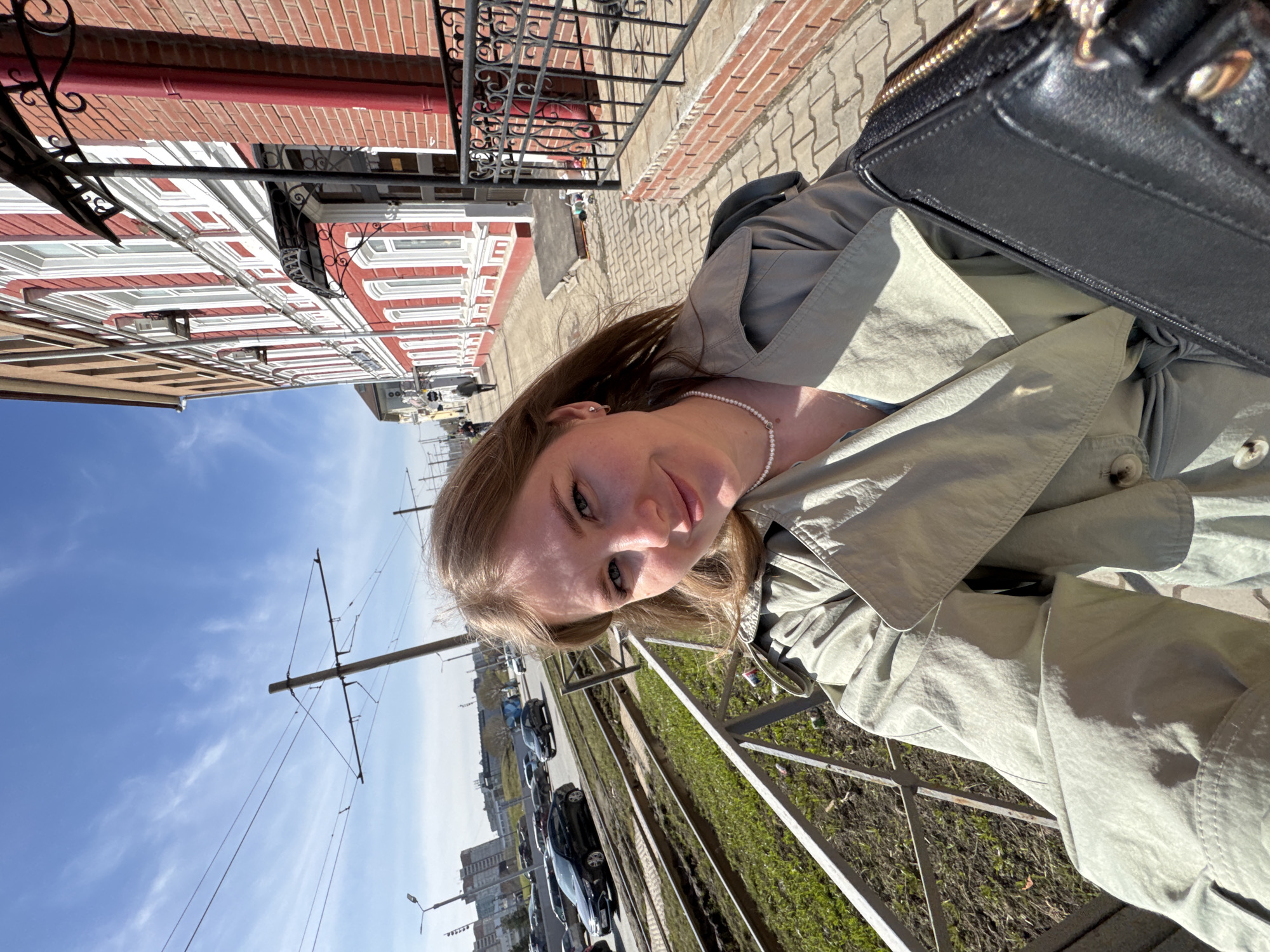 The image shows a woman in a light-colored trench coat and a pearl necklace, partially obscured by a black purse. She is on a street in Perm, Russia. The background features a row of buildings with red and white facades, decorative wrought-iron elements, and a brick sidewalk. On the other side of the street is a metal fence bordering a grassy area, with parked cars and distant high-rise buildings visible. Overhead tram lines and poles suggest public transportation. The sky is blue with scattered clouds.
