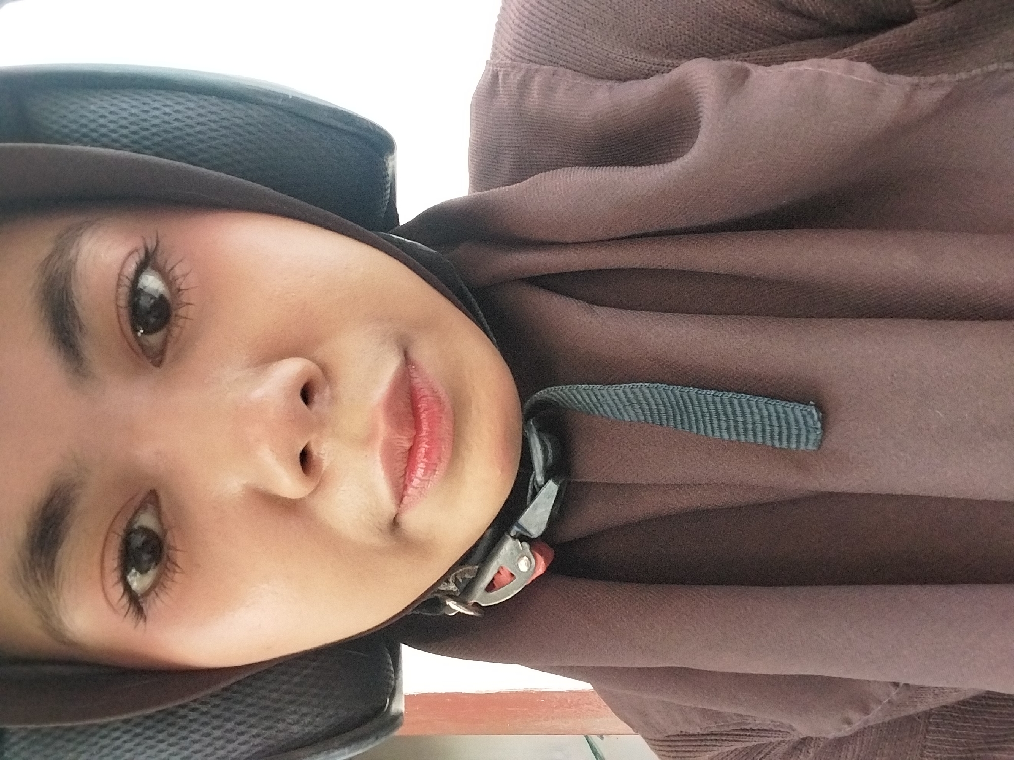 A close-up, head-and-shoulders shot shows a young woman wearing a dark brown hijab and a matching brown sweater. She is also wearing a black helmet with a mesh pattern on the sides, secured by a strap that is partially visible. The woman's face is the primary focus, with her eyes looking directly at the camera. Her eyebrows are neatly shaped, and her eyelashes are well-defined. She is wearing light makeup, including blush on her cheeks and lipstick on her lips. The background is blurred, but appears to be a building with a reddish-brown and white section on the left side of the frame, and a bright white area on the right. The image was taken in Tibojong, Indonesia.