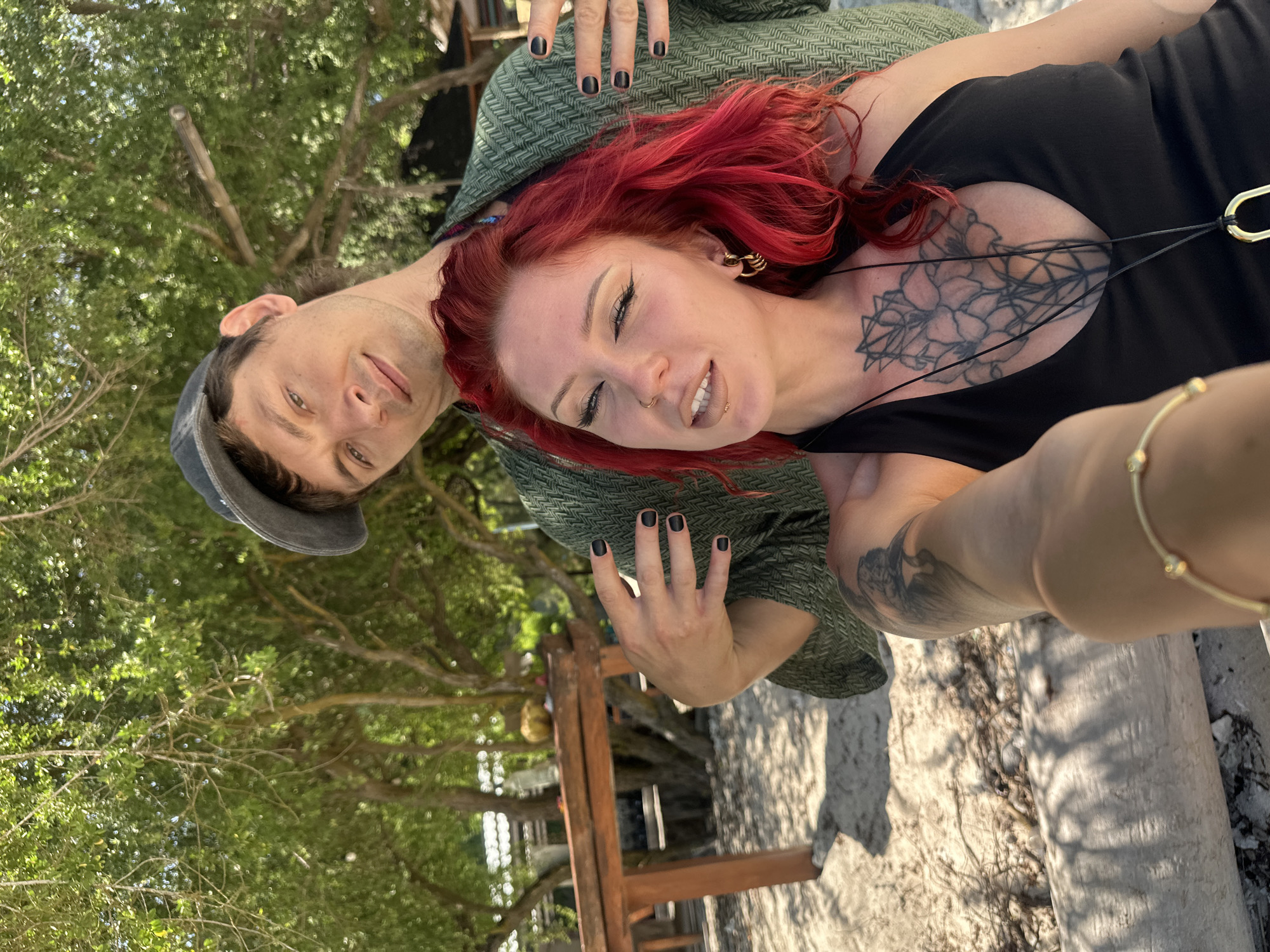 A selfie photograph shows a man and a woman with red hair, under a tree, on an outdoor surface in the city of Unknown, Indonesia. The woman, lying on the ground, is in the foreground and in focus, wearing a black top, visible tattoos, and facial piercings. The man is positioned behind her, partially out of focus, wearing a cap and a green patterned shirt or blanket draped over him. Both have manicured nails painted black. A gold bracelet adorns the arm closest to the camera. Wooden structures are visible in the background. The ground appears to be a light-colored surface, possibly sand or a similar material. The environment is filled with green foliage, suggesting a natural outdoor setting.