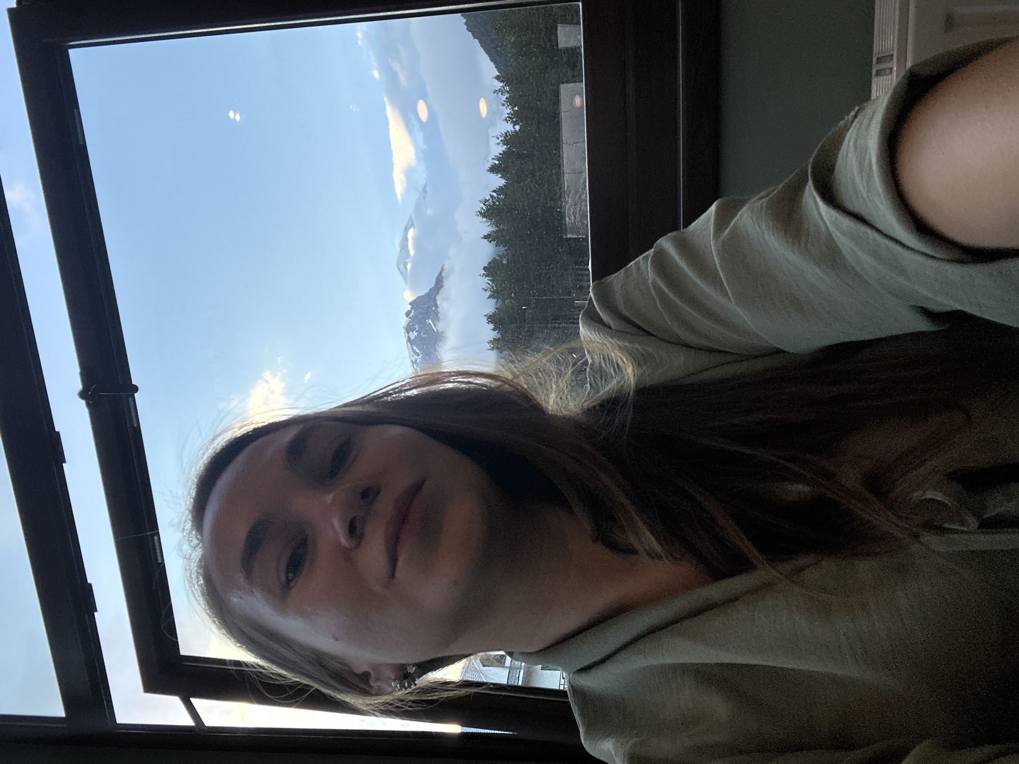 An indoor selfie features a young woman with long, brown hair, wearing a light green collared shirt. She is positioned in the foreground, looking towards the camera with a neutral expression. Behind her, a large window offers a view of a mountain landscape. The mountains, partially covered in snow and clouds, rise above a dense forest of evergreen trees. The scene appears to be set during daylight hours, likely in the late afternoon or early evening, given the soft lighting. The window frame and interior furnishings suggest a room with a view, possibly a restaurant or accommodation. The specific location is identified as Terskol, Russia, which is a well-known mountain resort area in the Elbrus region.