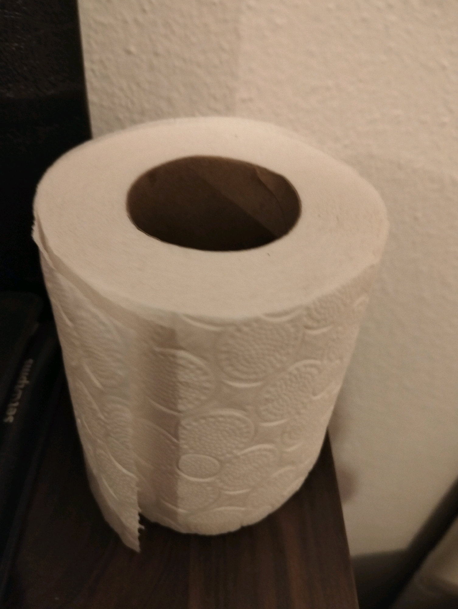 A roll of toilet paper is positioned on a dark brown wooden surface, likely a shelf or table, against a textured, light-colored wall. The toilet paper roll is white and features a circular pattern embossed on its surface. The core of the roll is visible and appears to be made of cardboard. To the left of the toilet paper, a dark, textured object, possibly a piece of furniture or electronics, is partially visible. The lighting in the scene is dim, casting soft shadows. The image was captured in Balçova, Turkey.