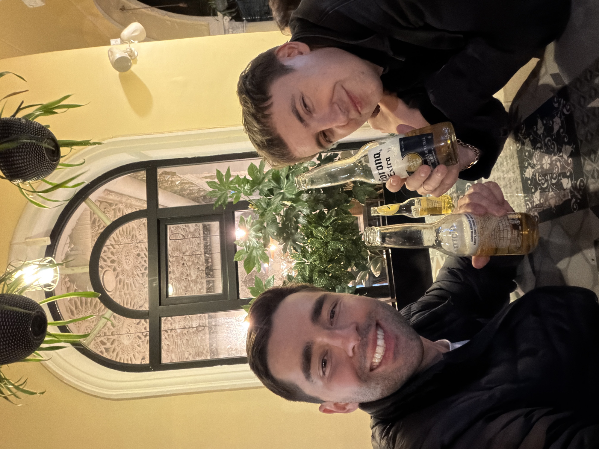 Two men pose indoors, holding Corona Extra beer bottles. They are in an establishment with a window featuring ornate designs. The image may be in the city of Batumi, Georgia. The scene includes plants, and a ceiling with a light fixture. The men are smiling at the camera.
