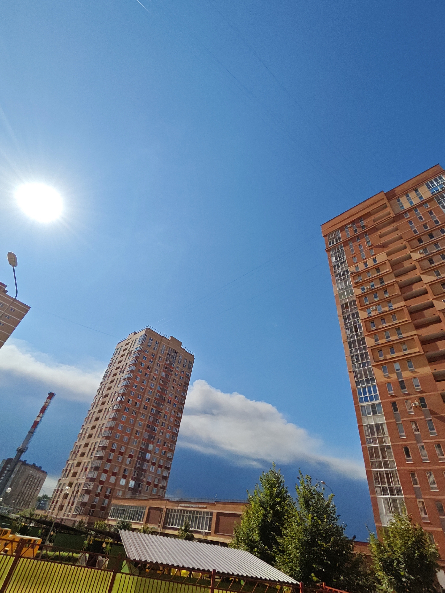 The image captures a bright, sunny day with a clear blue sky in the city of Shcherbinka, Russia. The sun is prominently visible in the upper left quadrant, casting a bright glare. Two tall, modern apartment buildings dominate the scene, both constructed with reddish-orange brick. The building on the right is closer to the viewer and features numerous balconies and large, multi-paned windows. The building on the left, further in the distance, is also tall and appears to have a similar architectural style. Between the two buildings, a construction crane with red and white markings is visible. In the foreground, there is a low-rise building with a modern, light-colored roof, surrounded by green trees and a yellow metal fence. The overall impression is of a residential area on a clear, fair-weather day.