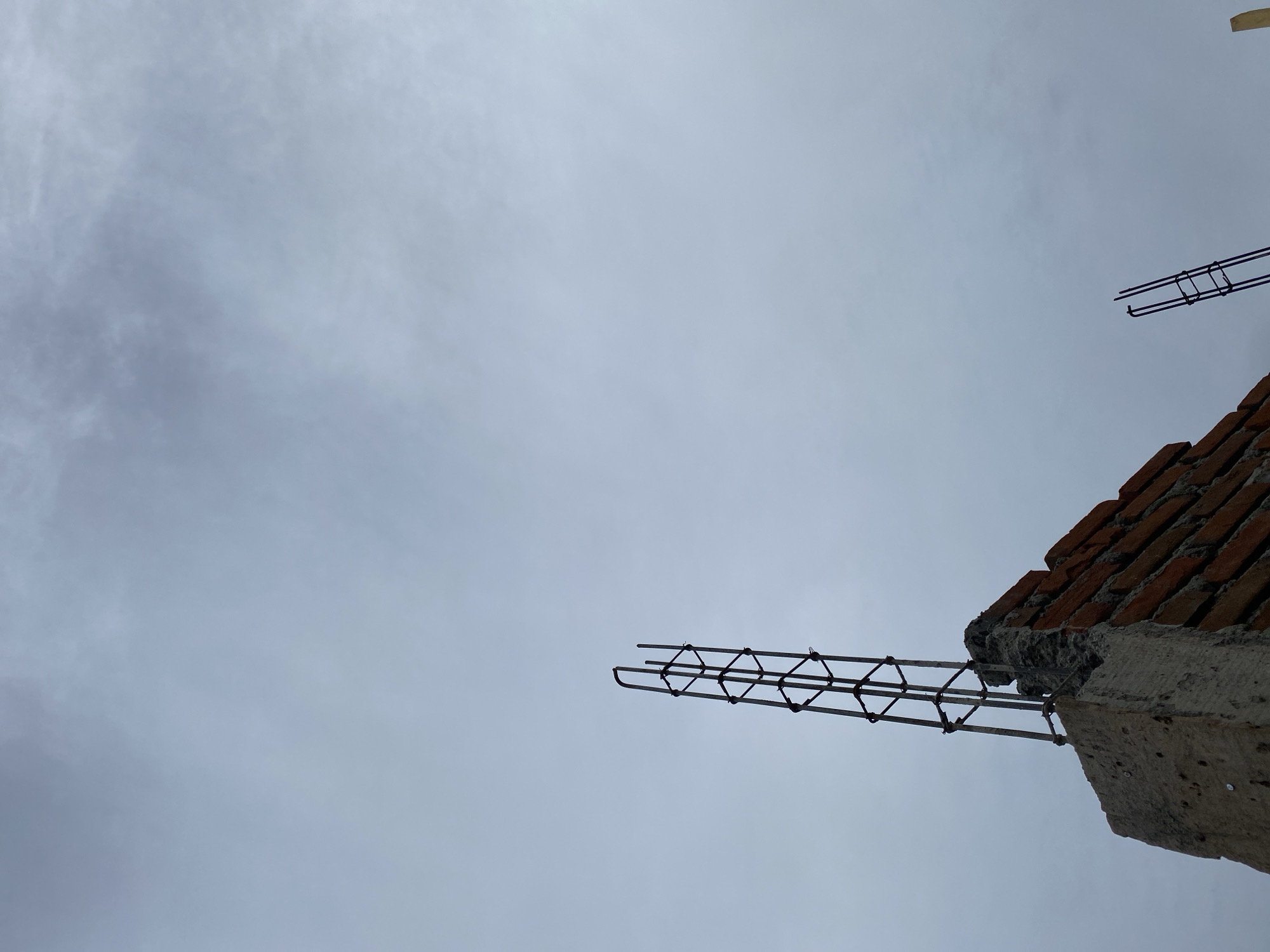 An overcast sky provides a muted backdrop for a construction site in Banda Aceh, Indonesia. In the foreground, the top of a brick wall, partially covered in concrete, juts upwards. Emerging from this structure is a metal rebar cage, a common element in reinforced concrete construction. Another, smaller rebar cage is visible to the right, also silhouetted against the sky. The scene suggests an ongoing building project, likely in its early stages. The cloudy weather contributes to a somber and industrial atmosphere.