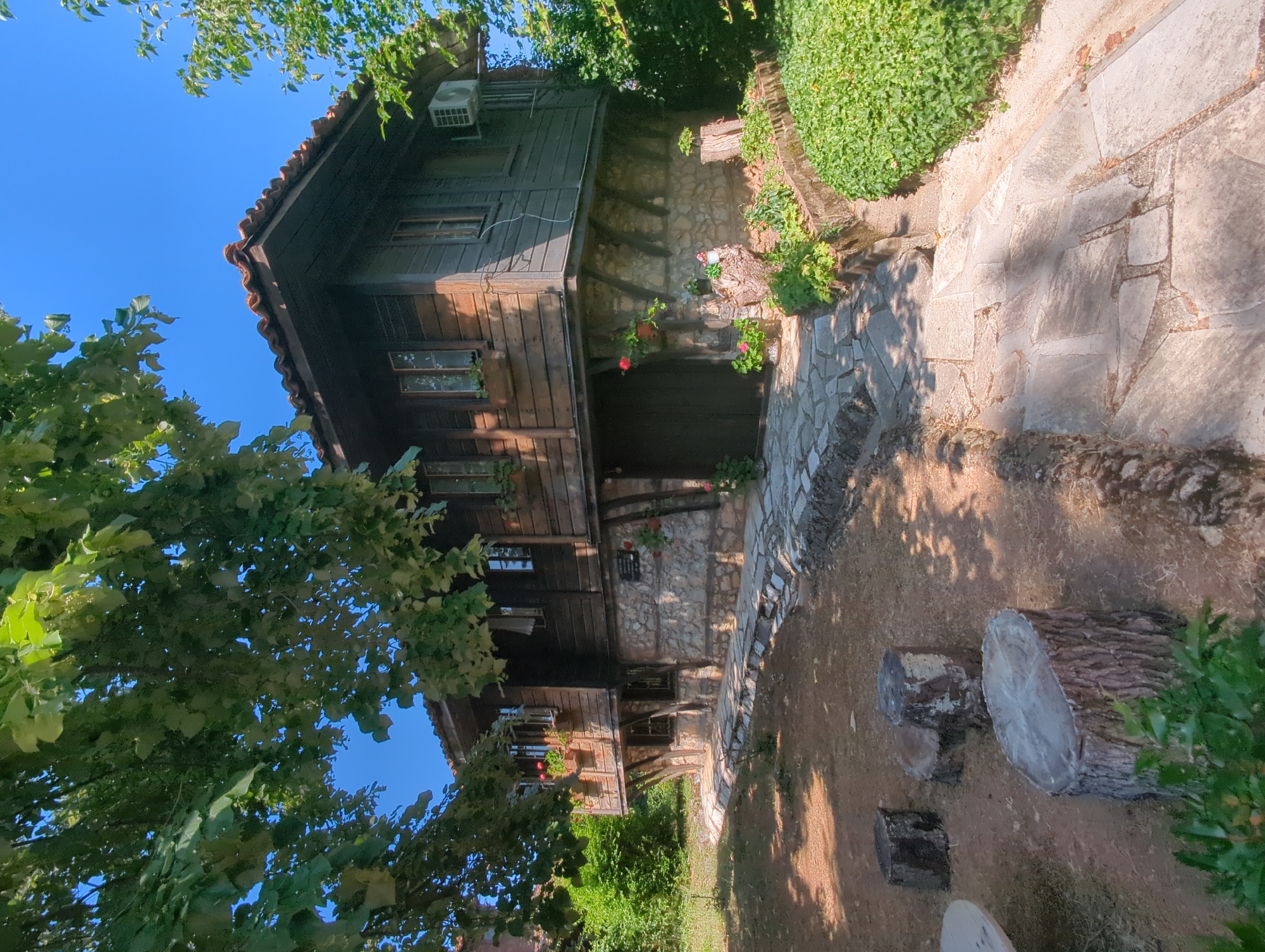 The image shows the exterior of a traditional wooden house in Malko Tarnovo, Bulgaria. The house has a stone foundation and a tiled roof. The wooden walls are weathered and dark, contrasting with the lighter stone base. Several windows are visible on the upper floor, some with flower boxes. A large, dark wooden door is present on the ground floor. The house is surrounded by greenery, with trees and bushes in the foreground and background. A stone pathway leads up to the house. Several tree stumps are scattered in the yard in front of the house, serving as seating. An air conditioning unit is mounted on the exterior wall of the upper floor. The sky is clear and blue.