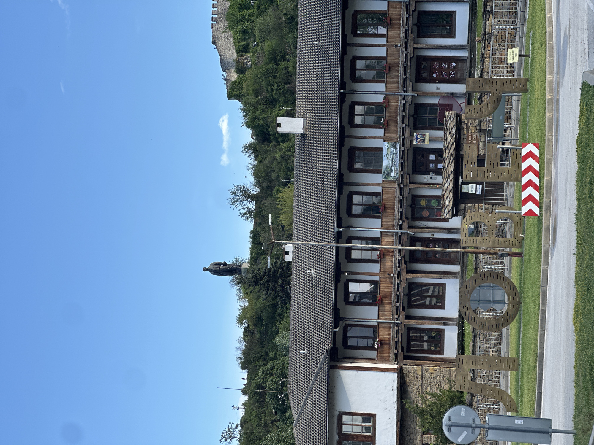 The image captures a street view in Lovech, Bulgaria, featuring a traditional Balkan-style building with a dark tile roof and multiple rows of windows, some with flower boxes. A large metal sculpture spelling "Lovech" stands in front of the building. A statue of Vasil Levski is perched on a hill above the building, and the Lovech Fortress can be seen further up the hill. A red and white directional sign is visible near the road, along with a road sign. The sky is clear blue, and the scene is bathed in daylight.