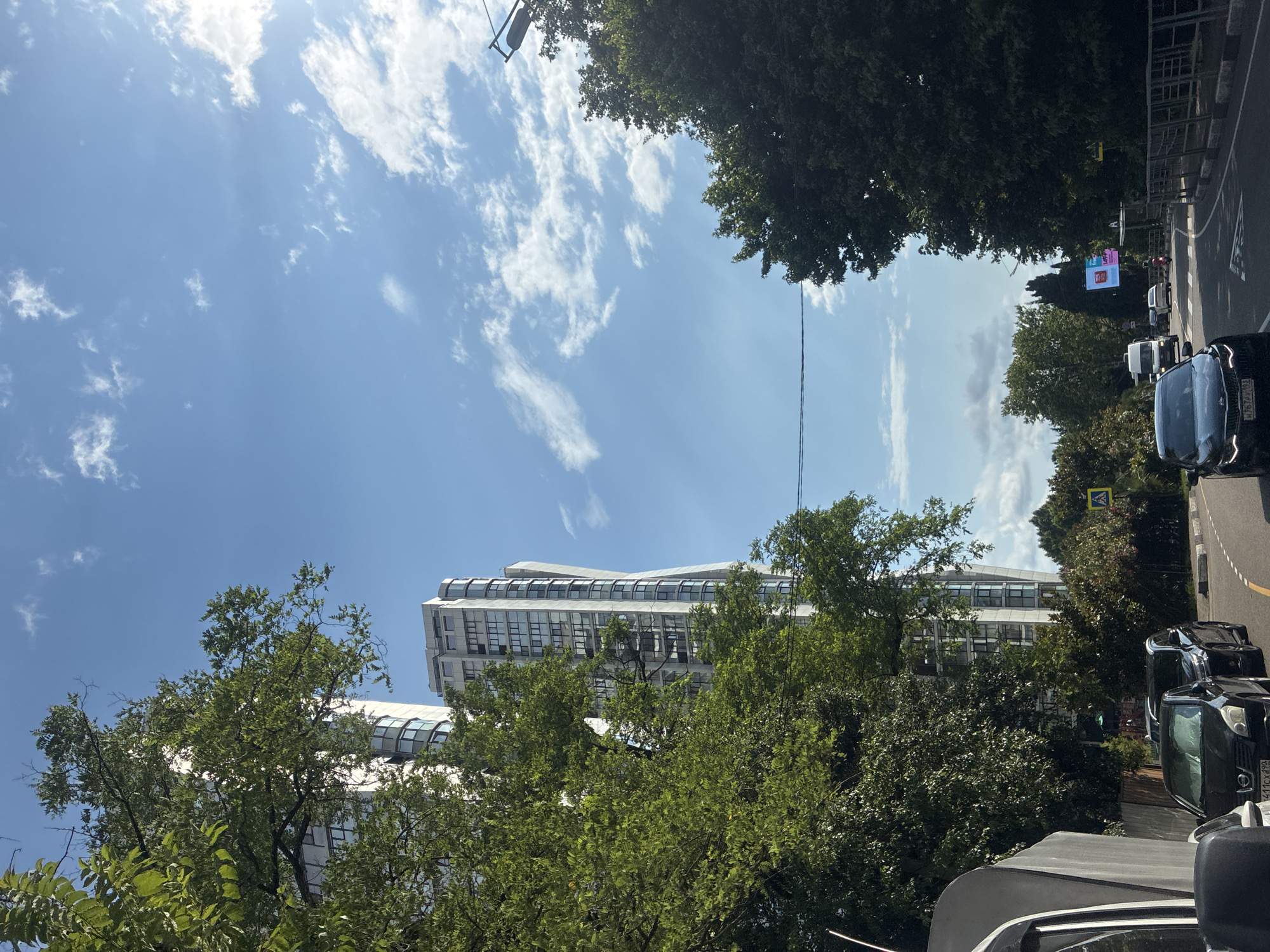 The image shows a street scene in Sochi, Russia, on a sunny day with scattered clouds. The perspective is looking up towards the sky, with lush green trees dominating the foreground. A modern, multi-story building with a unique, curved architectural design is visible behind the trees.
Several cars are parked or driving on the right side of the street, which curves away from the viewer. The cars appear to be modern vehicles. A digital billboard is visible in the distance, displaying bright advertisements. There is a pedestrian crossing sign visible on the side of the road.
The time of day appears to be daytime, likely late morning or early afternoon, given the bright sunlight and clear sky. The weather is pleasant, suitable for outdoor activities. The presence of trees and the general urban setting suggest this is a developed area within the city. No people are clearly visible in the image.
Visible text includes what appears to be a license plate on the black car in the foreground which reads "477CX 190". The digital billboard also contains text and graphics, but they are not clearly legible.