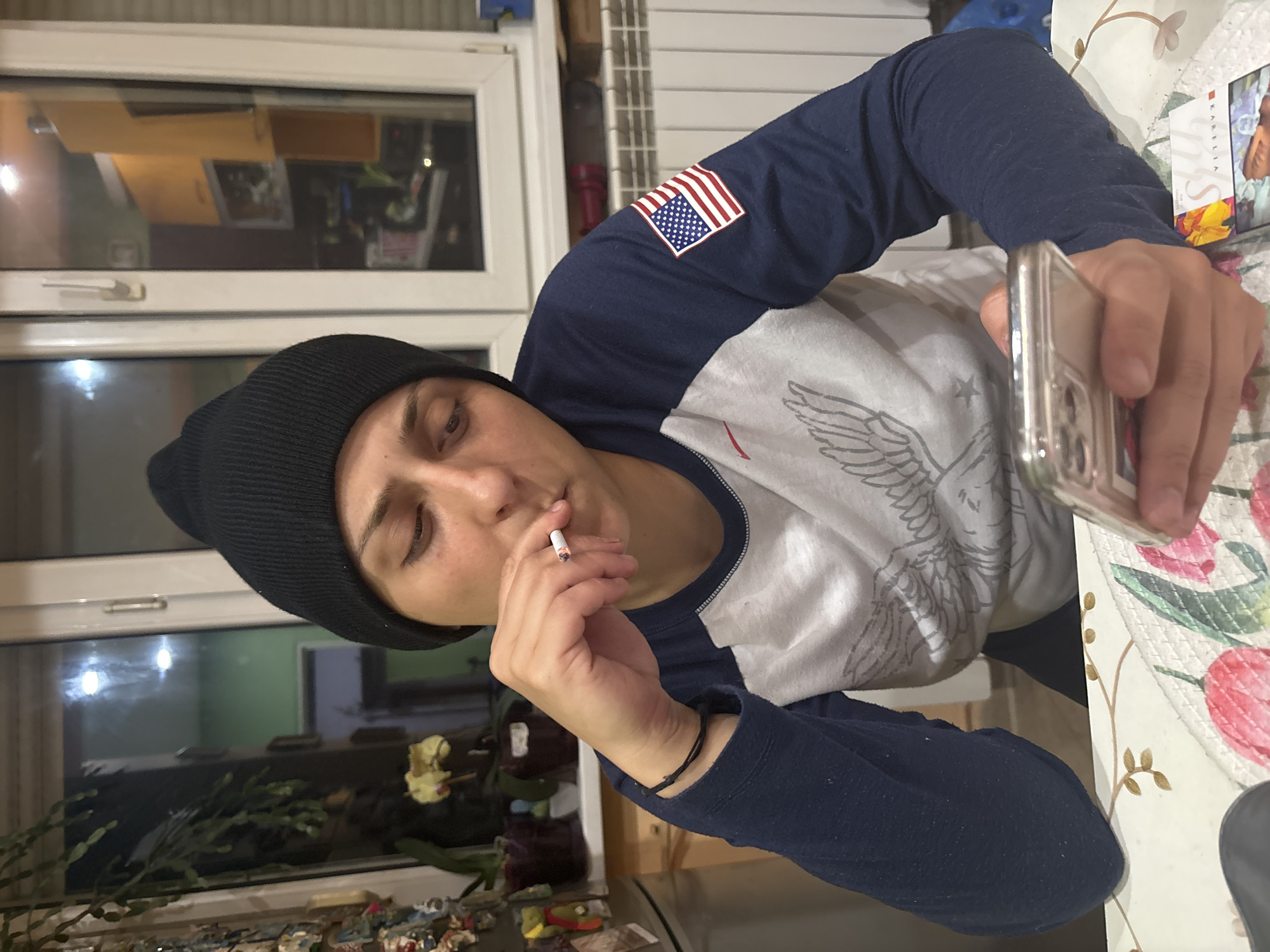 A person wearing a black beanie, a navy and white long-sleeved shirt with an American flag patch on the left sleeve and an eagle design on the torso, is holding a lit cigarette in one hand and a smartphone in the other. The person is sitting at a table with a floral-patterned tablecloth. The background shows a window with a reflection and plants near the window. The location cannot be ascertained from the image alone as being in Sofia, Bulgaria.