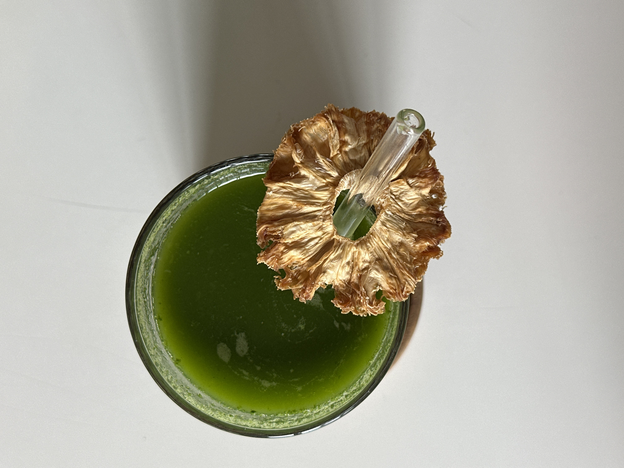 An overhead shot depicts a glass of green juice. The juice is topped with a dried pineapple ring and a glass straw. The glass sits on a white surface.
