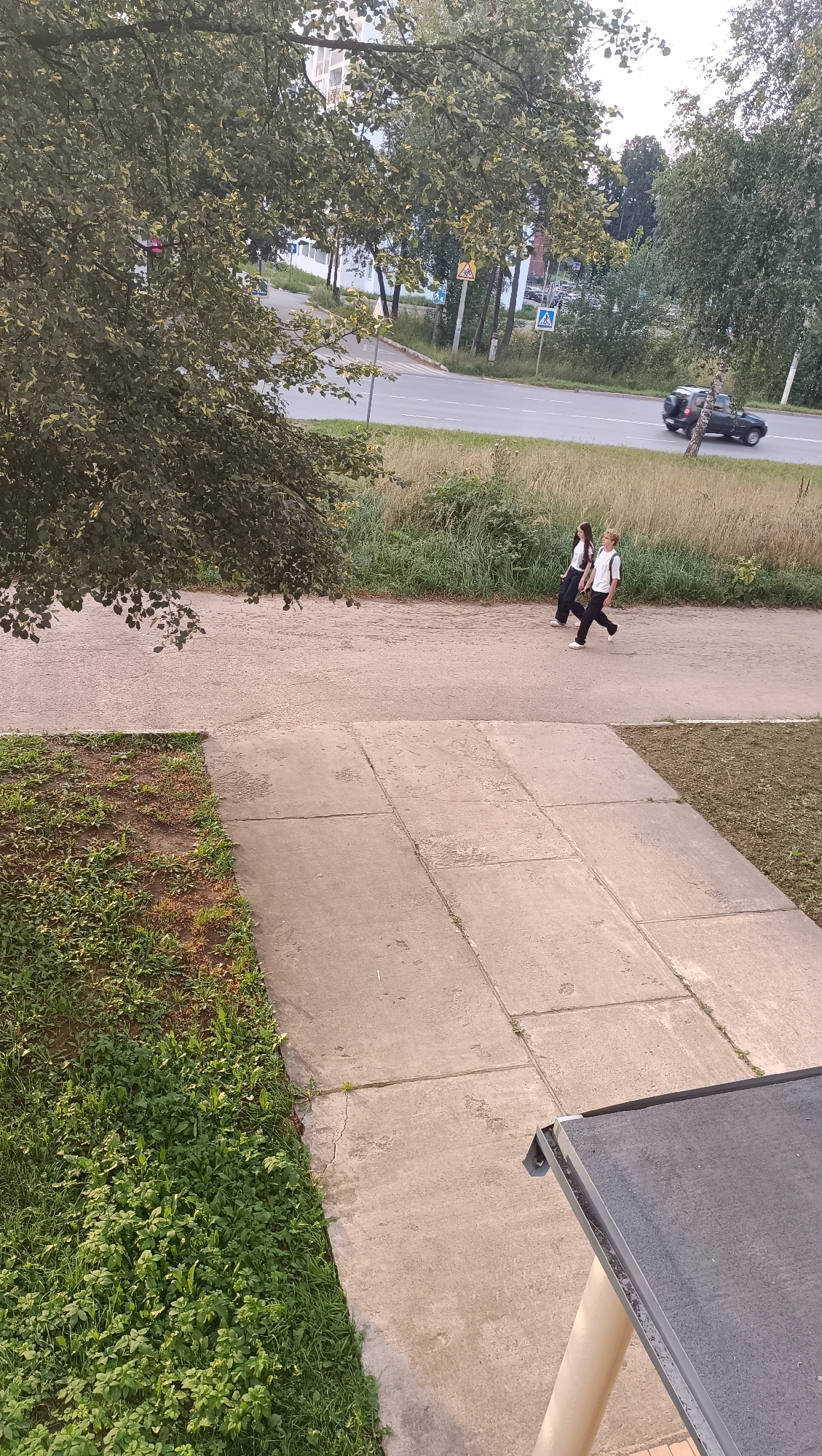 The image depicts a street scene with two young people walking on a sidewalk. The person on the left is wearing a white shirt and dark pants, while the person on the right has blond hair and is wearing a white shirt and dark pants with a backpack. They are walking away from the camera on a paved path. Behind them, a road is visible with a dark-colored SUV driving on it. The area is lined with grass and trees, suggesting a park or suburban environment. Road signs, including a pedestrian crossing sign and a directional sign, are also visible along the road. The sky is overcast. This scene is consistent with an outdoor location in Chaikovsky, Russia.