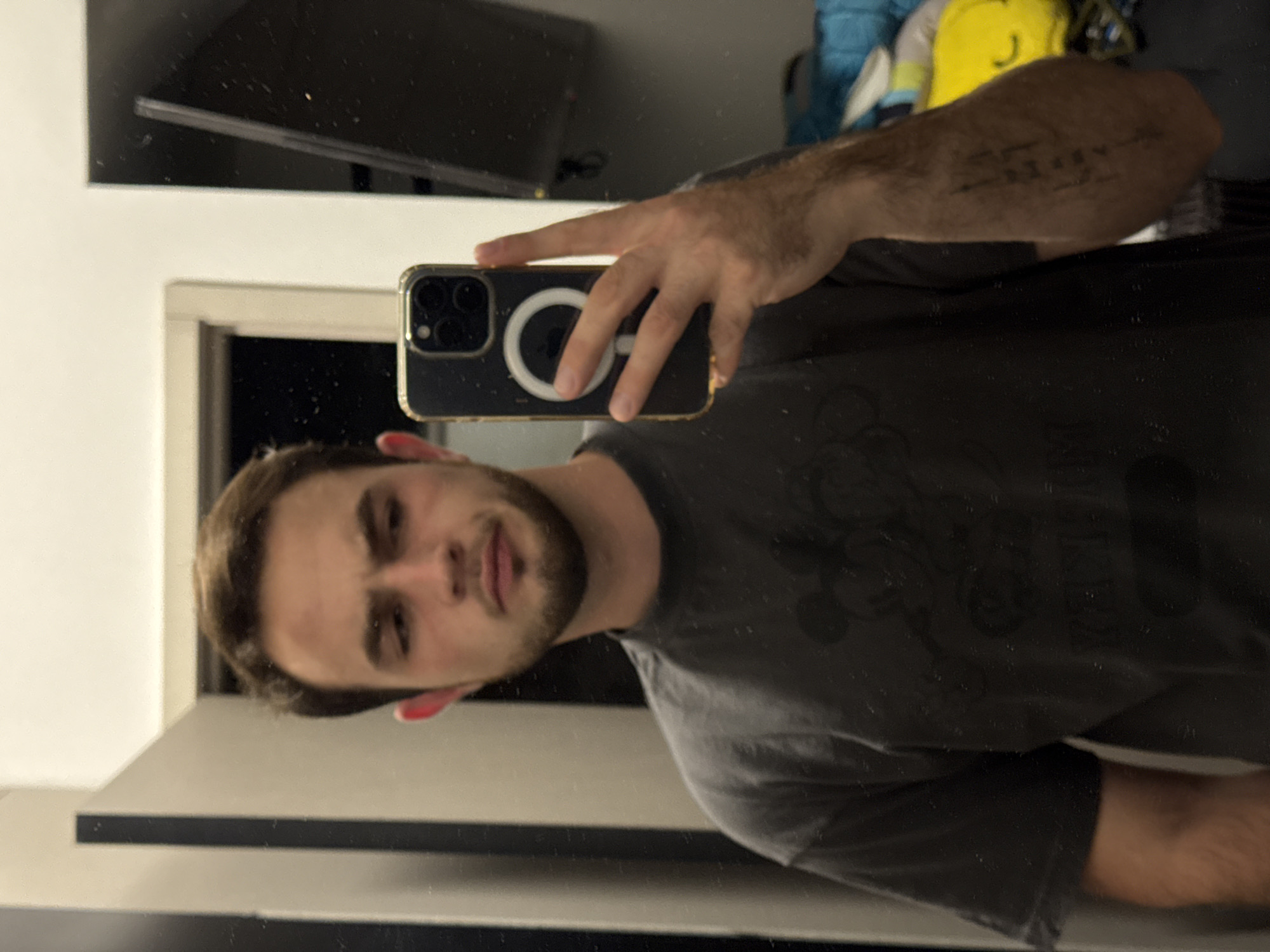 A man is taking a selfie in a mirror. He is holding a smartphone with a magnetic charging ring on the back. The reflection shows a room with a dark TV screen mounted on the wall. A doorway can be seen in the background. The man has short brown hair, a beard, and is wearing a gray T-shirt. There is a tattoo on his forearm. The location is not identifiable as San Bernardino Tlaxcalancingo, Mexico.