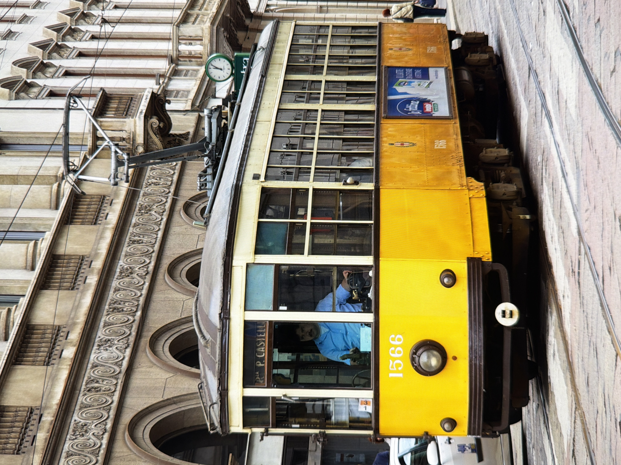 The image depicts a yellow tram with the number "1566" prominently displayed on its front. A man wearing a blue shirt is visible in the driver's compartment. The tram is on a street with cobblestone paving and tram tracks. In the background, there is a building with an ornate facade, featuring arches, columns, and decorative friezes. A woman is seen walking away from the tram on the right side of the image. A clock with a green face and white numbers is mounted on the building above the tram's roof. The scene appears to be an urban environment with architectural elements suggesting a European city. The provided information stating the location is Kyiv, Ukraine, is not supported by visual cues within the image itself.