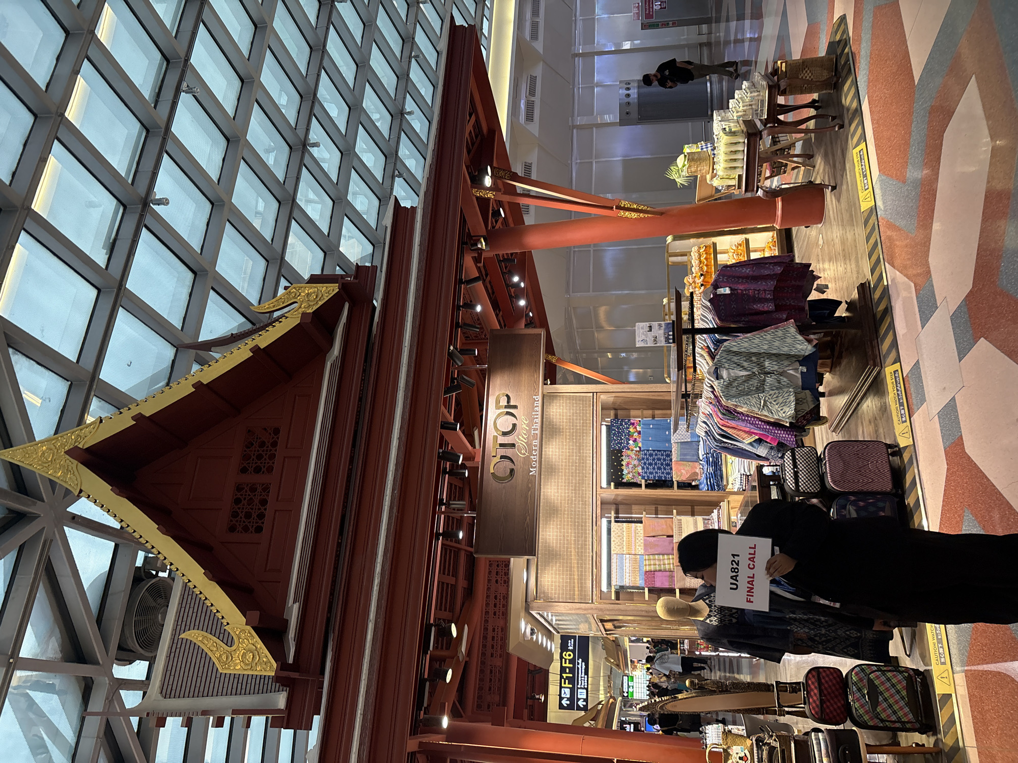 In an indoor marketplace with a modern glass and metal ceiling structure, a person in a dark suit holds up a sign reading "UA821 FINAL CALL" in front of a clothing stall. The stall, designed with traditional Thai architectural elements like a red wooden roof with golden trim, displays various fabrics and garments. To the left of the stall, suitcases are stacked, and a mannequin is dressed in traditional attire. In the background, other shoppers walk through the brightly lit space, and directional signs indicate "F1-F6." The floor features a patterned design of red, pink, and gray geometric shapes.