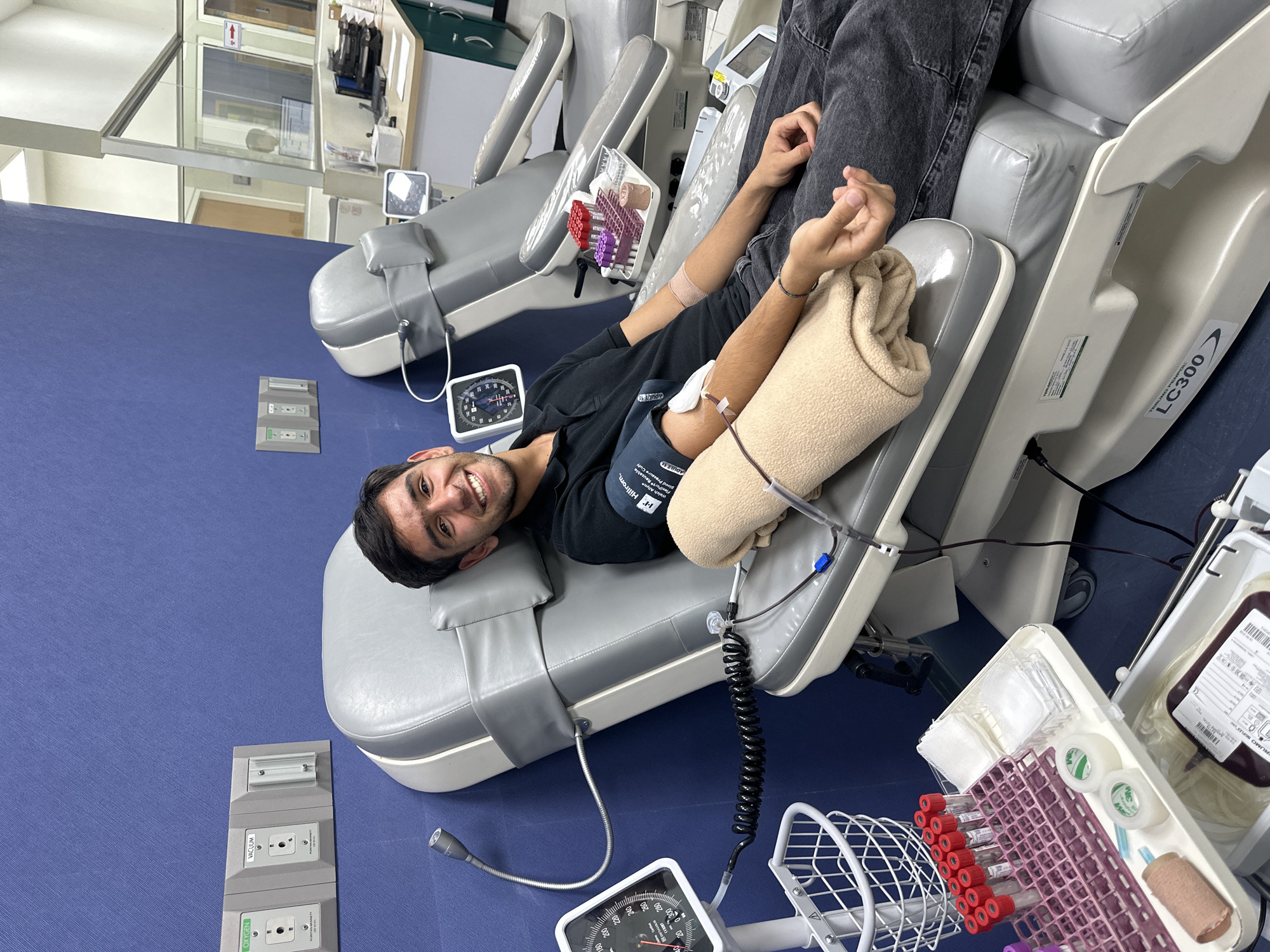 The image shows a man lying on a medical chair, apparently donating blood. He is smiling and facing the camera. A blood pressure cuff is on his arm, and a blood bag is nearby. Test tubes and medical supplies are positioned close to him, likely for the blood collection process. The setting appears to be a medical facility or blood donation center, with blue wall and other medical chairs in the background. There's equipment visible like blood pressure monitors, and various wall-mounted medical outlets. The text on the chair indicates its model is "LC300". A blood pressure cuff has the inscription "PR Hillrom." and "Welch Allyne." on the wall's oxygen outlet. The city of Huixquilucan de Degollado, Mexico cannot be determined from the provided image alone.