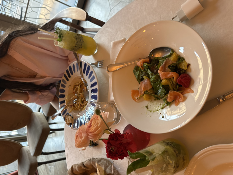 The image depicts a dining scene inside Casa Nostra restaurant in Yerevan, Armenia. The focal point is a white-clothed table laden with partially consumed meals. In the upper right, a person with long dark hair, wearing a pink collared shirt and a gold necklace, is partially visible, holding a mobile phone to their ear or side of the face.
On the table, a blue and white patterned bowl contains a small amount of leftover pasta, with a fork and spoon resting inside and a piece of bread on the rim. To its right, a tall glass holds a vibrant yellow-green drink with visible kiwi slices, mint leaves, and two white straws. Another clear glass on the left contains a green beverage with lime or kiwi slices and mint, featuring a wooden stirring stick. A white bread basket is positioned to the far left.
A white ceramic bowl in the foreground holds a salad composed of leafy greens, orange segments, slices of smoked salmon, and a red berry (likely a raspberry), with a silver spoon inside. Below this bowl, a silver-handled knife rests on the tablecloth. A white USB wall charger is visible on the tablecloth to the far right.
Table decorations include a small clear glass vase holding two pink ranunculus flowers and a small red vase containing one deep red ranunculus flower. The background shows two beige upholstered chairs with dark metal legs, and a large window offering a blurred view of an outdoor area, suggesting the restaurant is on an upper floor or has an expansive patio.