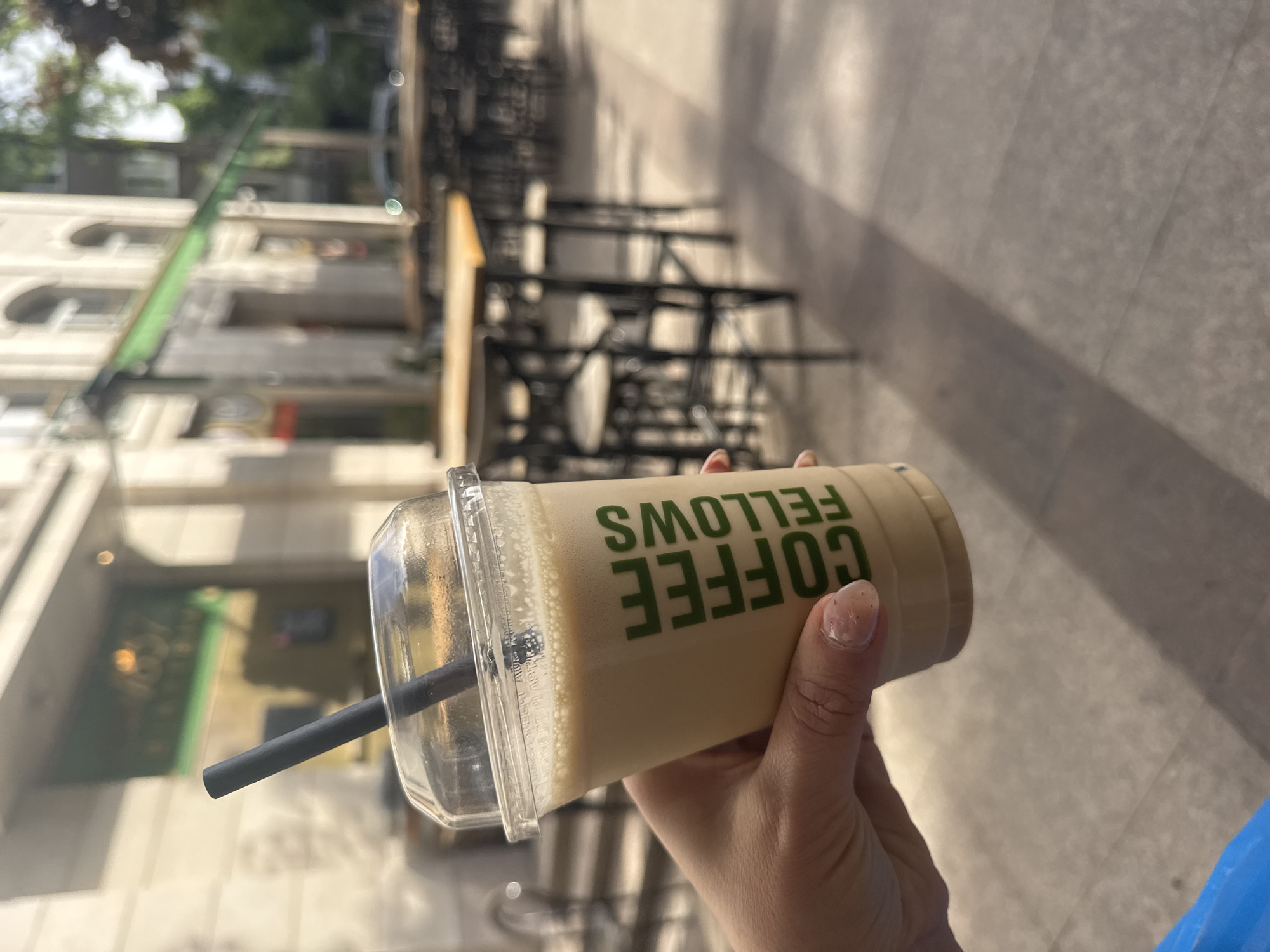 A person is holding a "Coffee Fellows" branded iced coffee in a clear plastic cup with a black straw. The drink appears to be a light tan color. The person's hand and part of their arm are visible. In the background, there is a blurred street scene with buildings, trees, and outdoor cafe seating. The photo was likely taken in Sofia, Bulgaria, as the Coffee Fellows brand is present in that city. The foreground is in focus while the background is intentionally blurred.