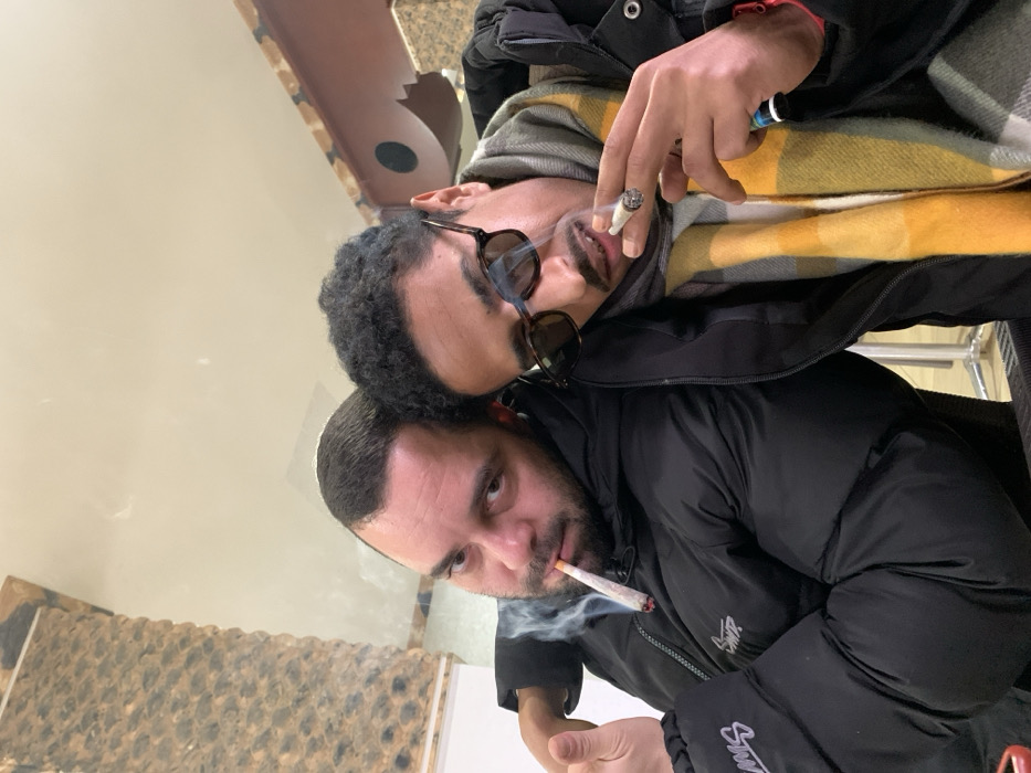 Two men are posing closely together. One, with a beard and short dark hair, holds a lit cigarette. The other, with curly dark hair and sunglasses, also holds a lit cigarette. Both are wearing dark jackets. The top man has a yellow and gray scarf. The background is blurry.