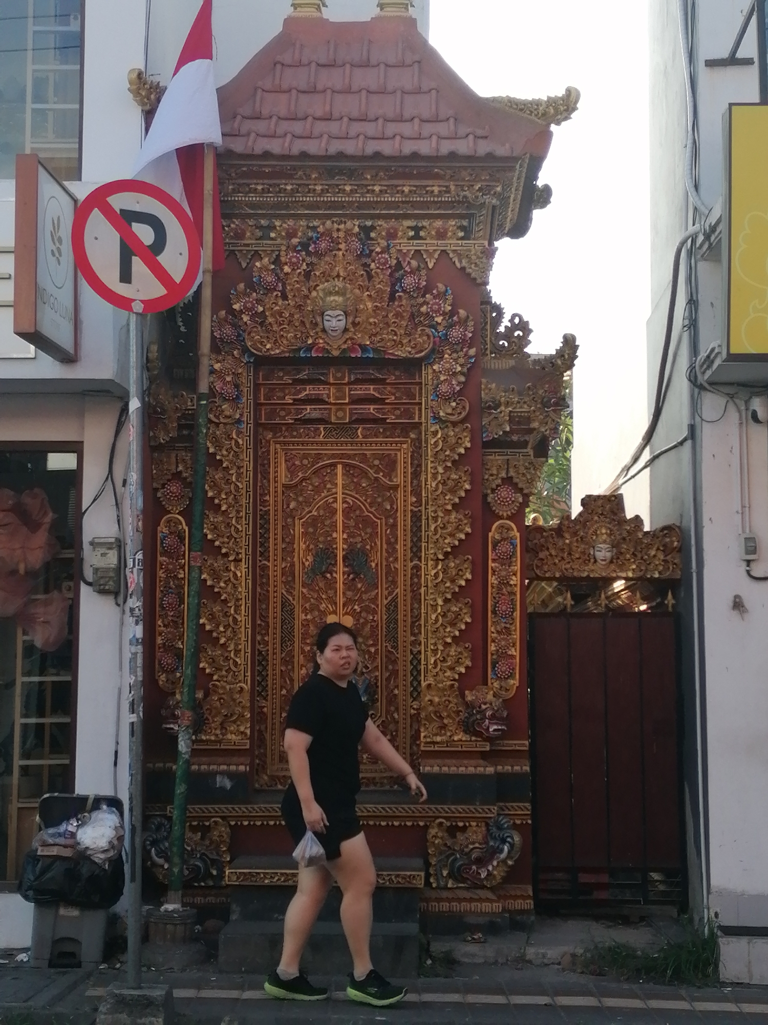In Ubud District, Indonesia, a woman walks past an ornate temple gate. She is wearing a black t-shirt and shorts, and holding a plastic bag. The temple gate is made of wood and decorated with intricate carvings and gold accents. A red and white flag is flying from a pole next to the gate. A "no parking" sign is visible on the same pole. In the background, there is a building and some electrical wires. A trash can is located to the left of the gate. The scene is illuminated by natural light, suggesting it is daytime.