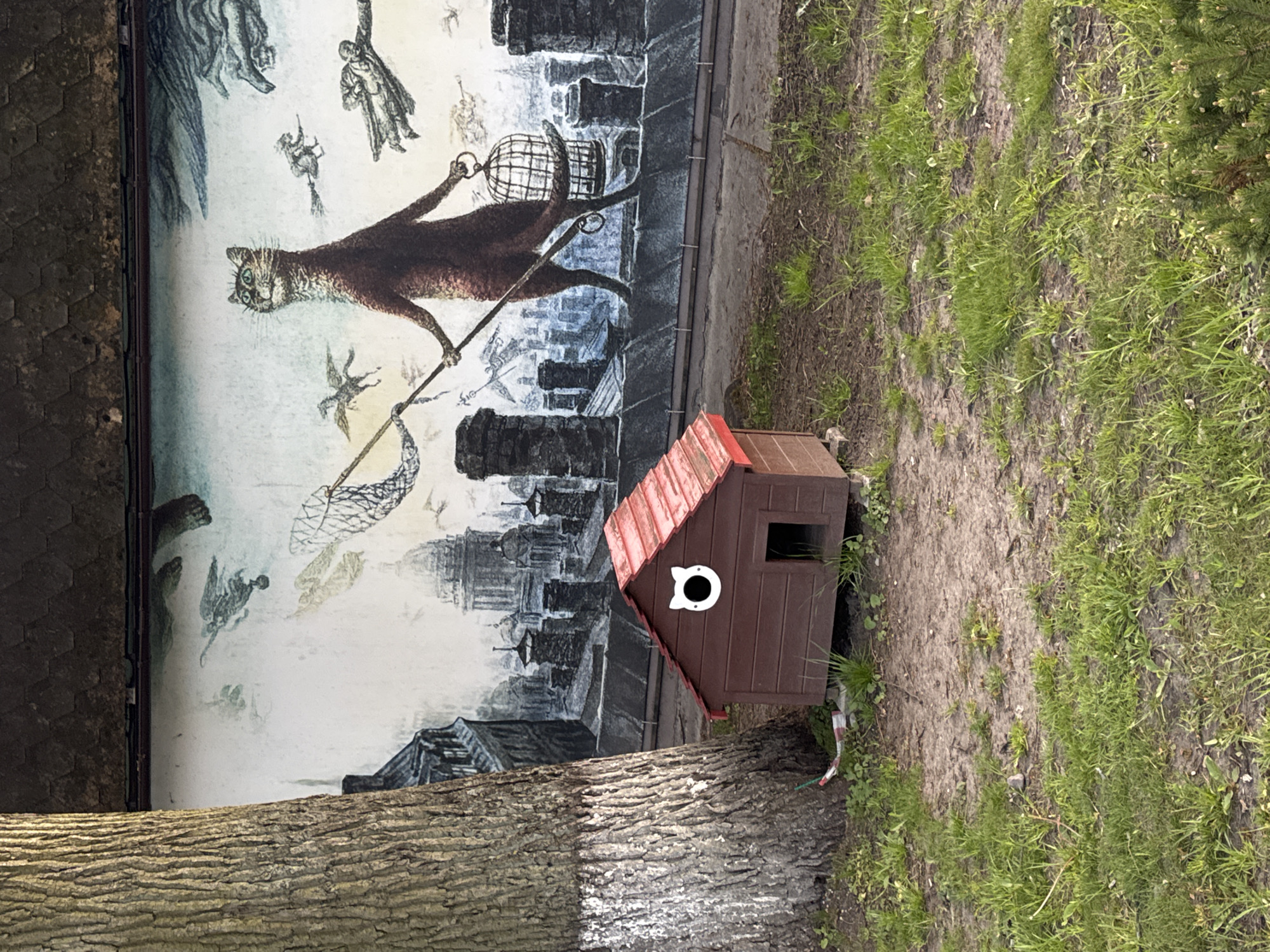 The image shows an outdoor scene with a mural and a small animal house. The mural depicts a large cat carrying a cage and a net, set against a backdrop of rooftops and flying figures. In front of the mural, partially obscured by the base of a tree, is a small, brown animal house with a red roof and a cat-shaped entrance hole. The surrounding area consists of dirt and patches of grass. Based on the mural style and setting, the location is likely in Zelenogradsk, Russia, known for its cat-themed street art.