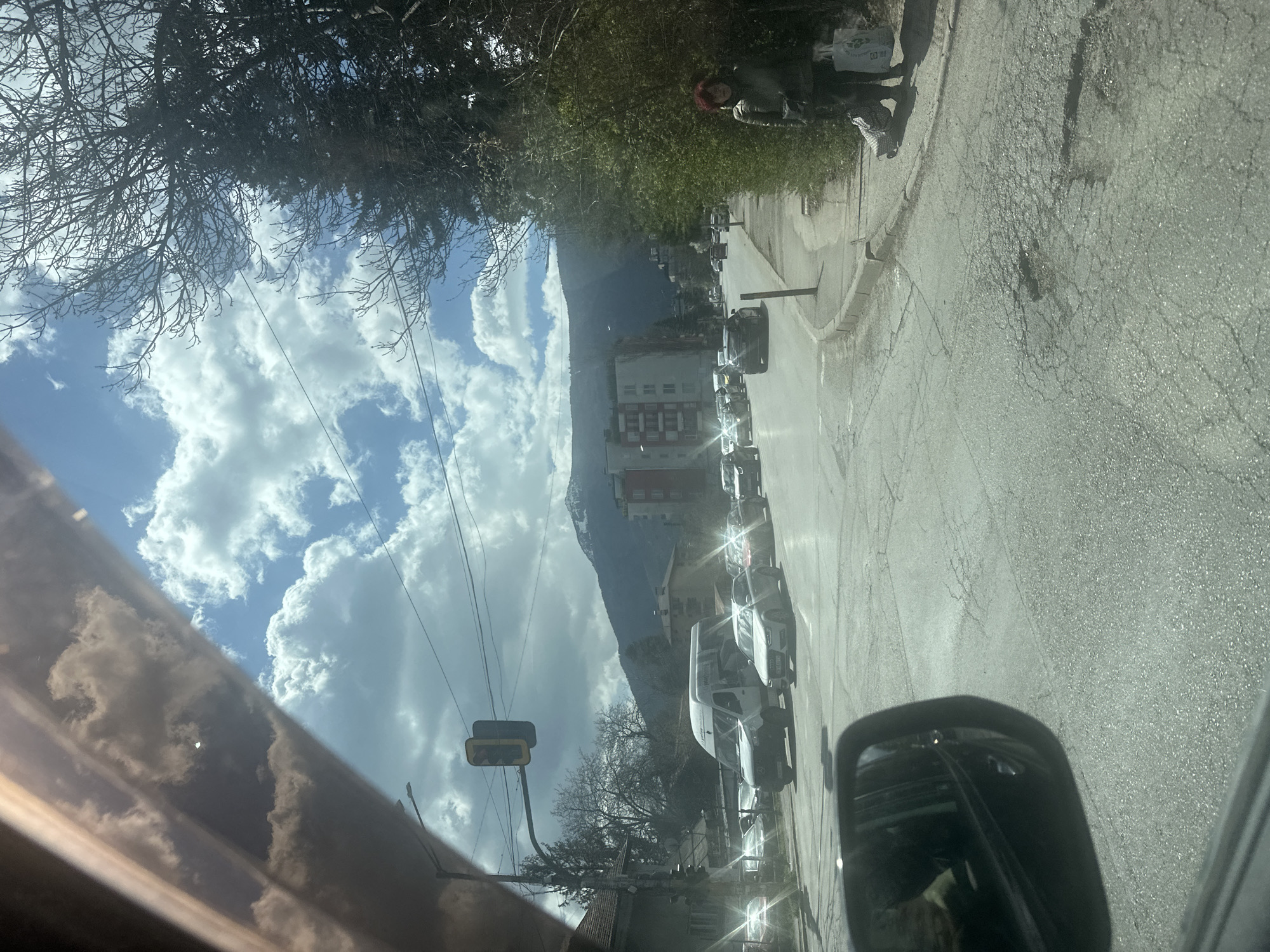 The image captures a street scene in Unknown, Unknown, taken from inside a vehicle. The view is dominated by a line of parked cars along the right side of the road, leading towards a tall building with a mountain backdrop. The sky is partly cloudy, visible above and behind the building. On the left, tree branches and power lines are seen against the sky. A traffic light is also visible on the left side of the road. In the upper right, a person with red hair is standing on the sidewalk. The side mirror of the vehicle is visible in the lower right corner. The overall scene depicts a typical urban environment with natural elements.