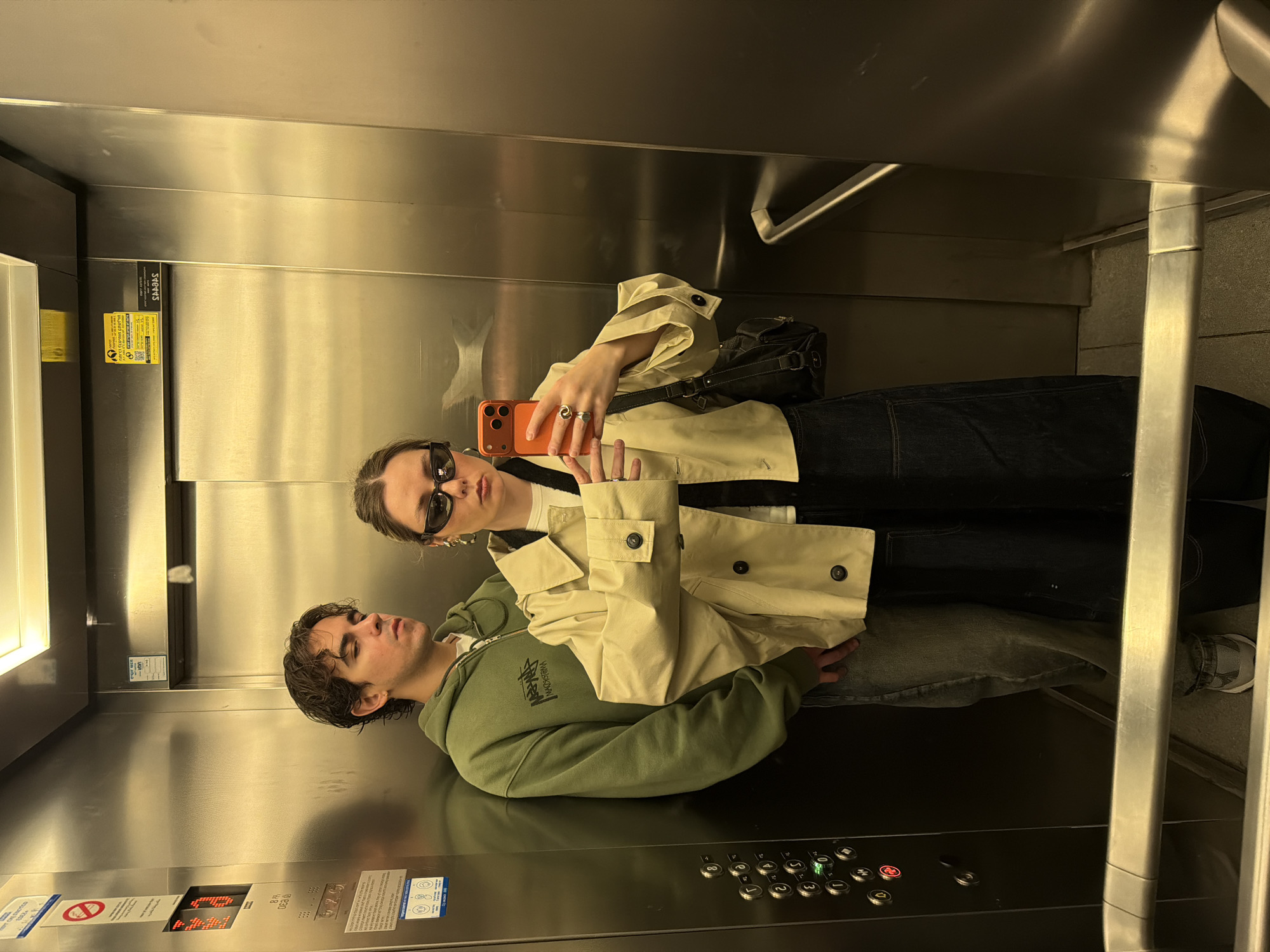 Two people are inside an elevator. The elevator's interior is stainless steel, with a panel of buttons indicating the floor level, and a digital display. The two individuals are facing the elevator door. One person, seemingly male, is lying on the elevator floor wearing an olive green hoodie. The other person, seemingly female, stands next to them taking a selfie with a red phone. She is wearing a beige trench coat and sunglasses. The location appears to be in Tel-Aviv, Israel, evidenced by the yellow 'Sparkes' label with a phone number starting with '03' and safety information printed in Hebrew on the elevator wall.