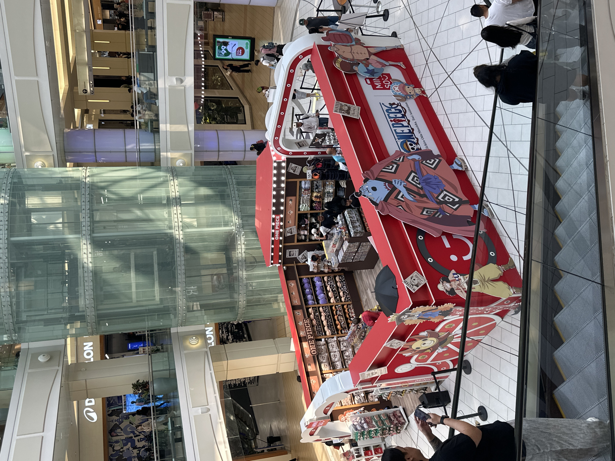 The image captures a bustling scene within a multi-level shopping mall, likely in Burnaby, Canada, given the presence of "D-LO" and "LOON" signage, which are associated with the Metropolis at Metrotown mall in that city. The central focus is a brightly colored, red and white kiosk themed around the popular anime and manga series "One Piece." The kiosk is adorned with large character cutouts and displays various merchandise, including hats and plush toys, arranged on shelves.
Several individuals are present. In the foreground, a person on an escalator is taking a photograph with their phone, capturing the scene. Another person in the foreground on the left is looking down at their phone, seemingly engaged with it. Further into the mall, on the upper levels, groups of people are seen browsing shops or walking. A prominent glass elevator shaft, a common architectural feature in modern malls, is visible to the left of the kiosk, reflecting the surrounding environment. The overall atmosphere suggests a commercial retail setting with ongoing visitor activity.
