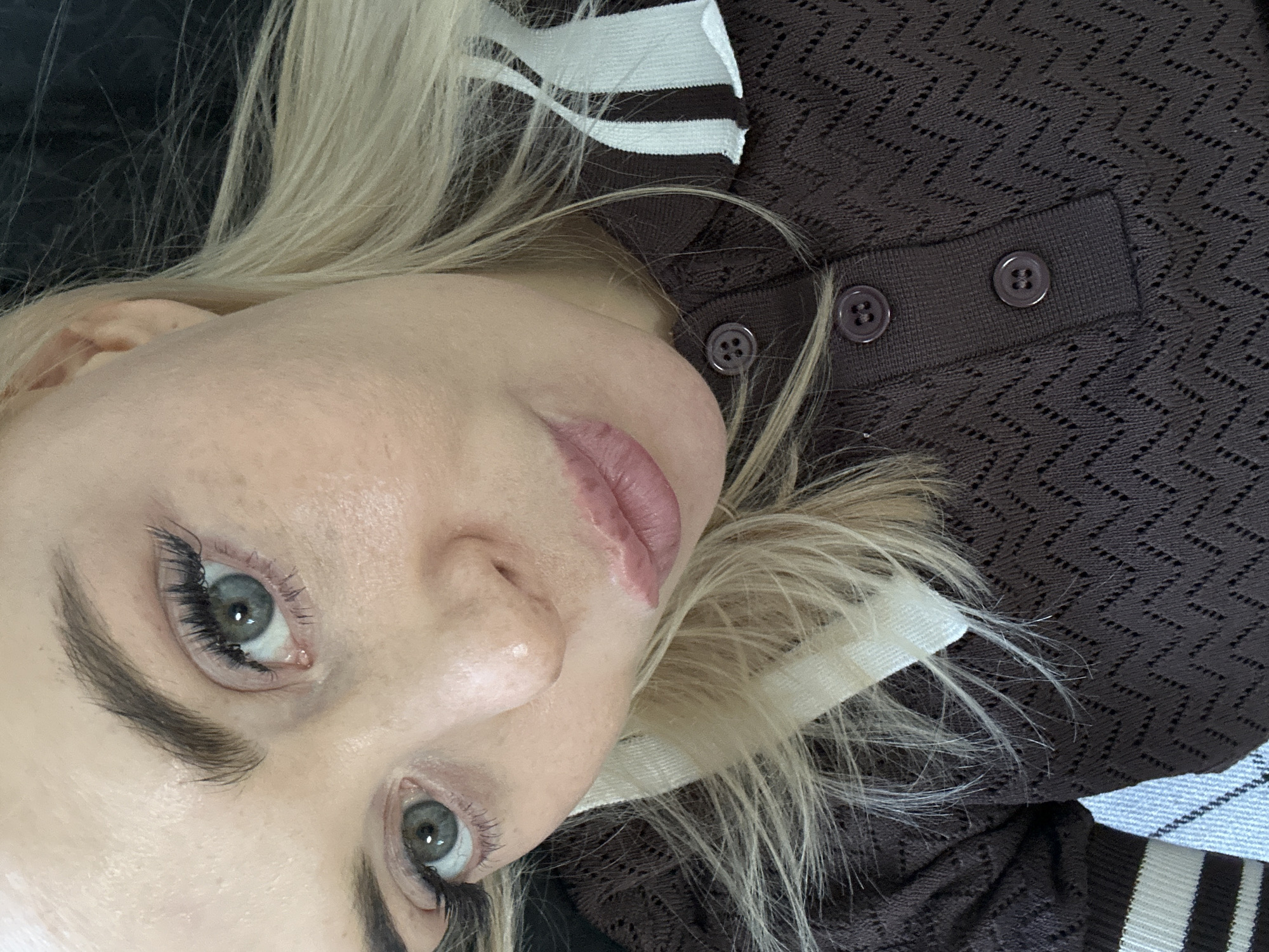 A close-up, rotated portrait shows a fair-skinned person, likely a woman, with long blonde hair and striking eyes, looking directly at the viewer. The image is oriented with the person's head tilted significantly, almost lying on their side.
The subject has bright, light blue or grey eyes, prominent dark eyelashes (possibly enhanced with mascara or extensions), and well-defined, dark eyebrows. Their lips are slightly parted, revealing a light pink or nude lipstick, with some visible texture on the upper lip. The skin appears clear with natural texture.
The person is wearing a dark brown or charcoal-colored garment, possibly a sweater or cardigan, which features a distinctive patterned knit, resembling a chevron or zig-zag design. The garment has a collar or trim with contrasting white and dark stripes, and dark brown, circular buttons are visible on the front placket. The blonde hair is fanned out around the head, blending into the dark, indistinct background. A faint glimpse of a lighter, possibly striped or textured fabric can be seen on the right side of the image, contributing to the soft background.
The overall scene is a posed portrait, focusing on the subject's face and upper body. There is no discernible activity beyond posing. No visible text is present. The setting appears to be indoors, though specific location cues are absent. The expression is neutral and direct.