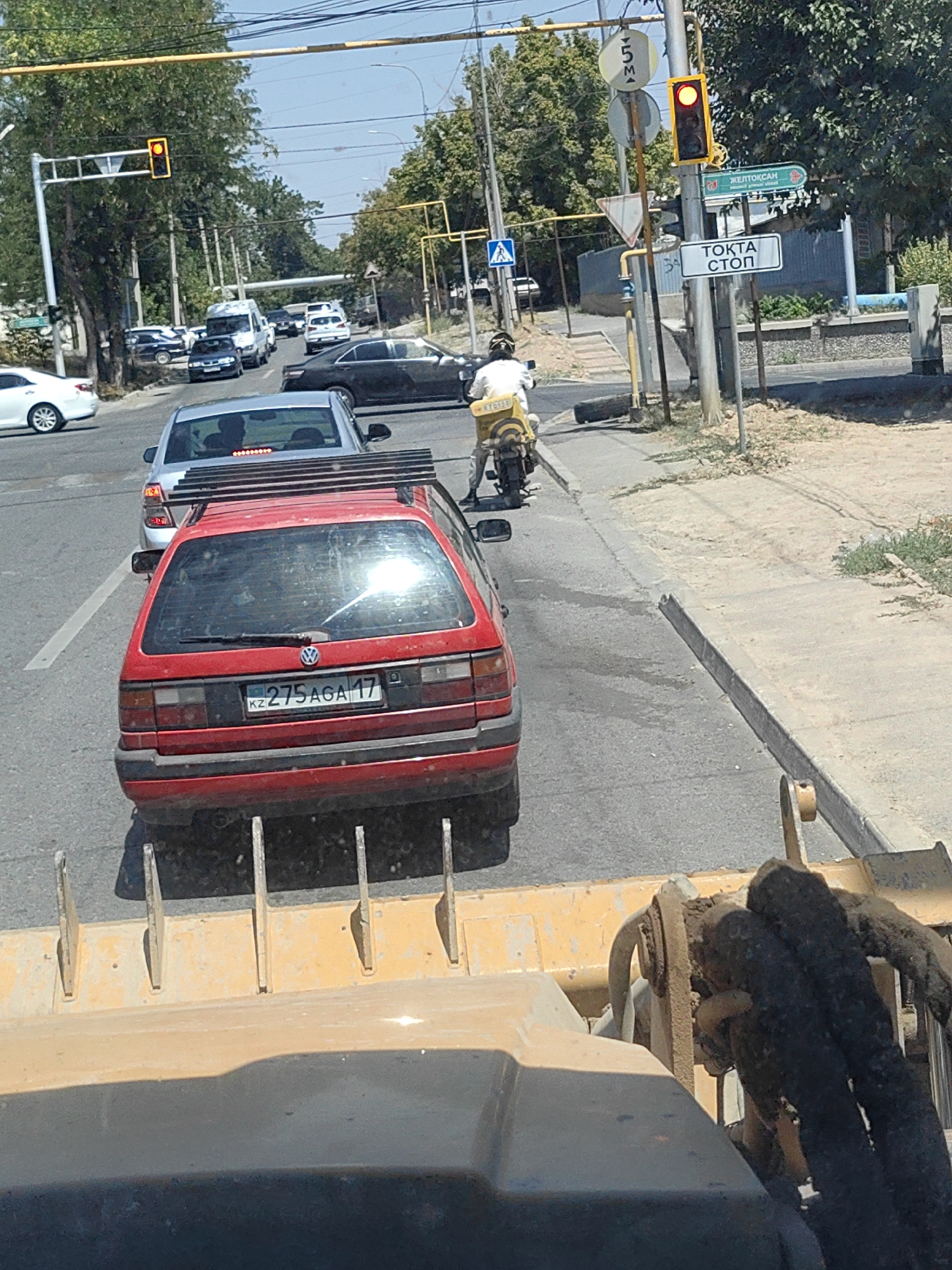 The image shows a street scene in Shymkent, Kazakhstan. A red Volkswagen hatchback is stopped in traffic, its license plate reading "KZ 275AGA 17". Behind it, a silver sedan is also in traffic. A person on a motorcycle, wearing a helmet and a yellow jacket, is stopped near a traffic light that is illuminated red. A street sign in the background reads "ТОҚТА СТОП", which translates to "STOP". Another sign indicates "ЖЕЛТОҚСАН", likely referring to a street name or area. The environment is an urban setting with trees lining the street and a clear blue sky. A construction vehicle or equipment is partially visible in the foreground.