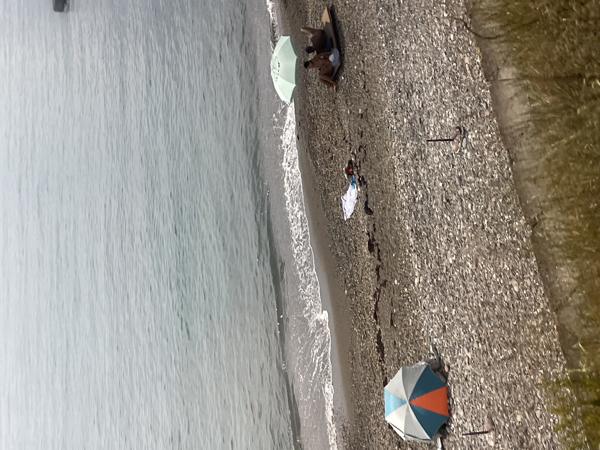 The image depicts a pebble beach scene in Sochi, Russia, with the Black Sea in the background. In the foreground, a colorful beach umbrella with orange, blue, and white panels is set up on the pebbled shore. Further down the beach, closer to the water, a light green beach umbrella is open, providing shade for two individuals. One person is lying down on a mat, while the other is seated. A towel is laid out on the pebbles near them, along with what appears to be a small pile of belongings. The sea is calm with gentle waves lapping at the shore. The overall atmosphere is one of relaxation and a summer day at the beach.